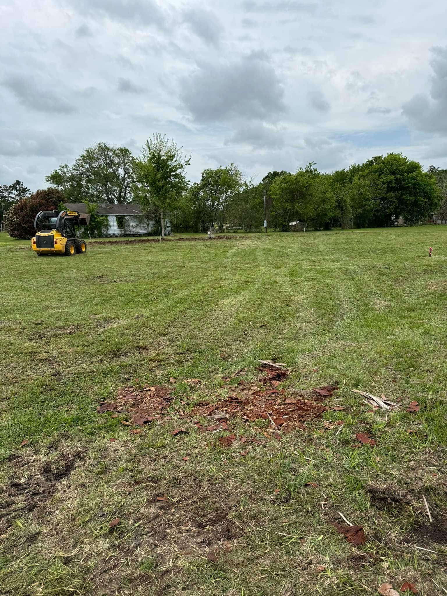 A small tractor working on a grassy field, trees and a house in the background under a cloudy sky.