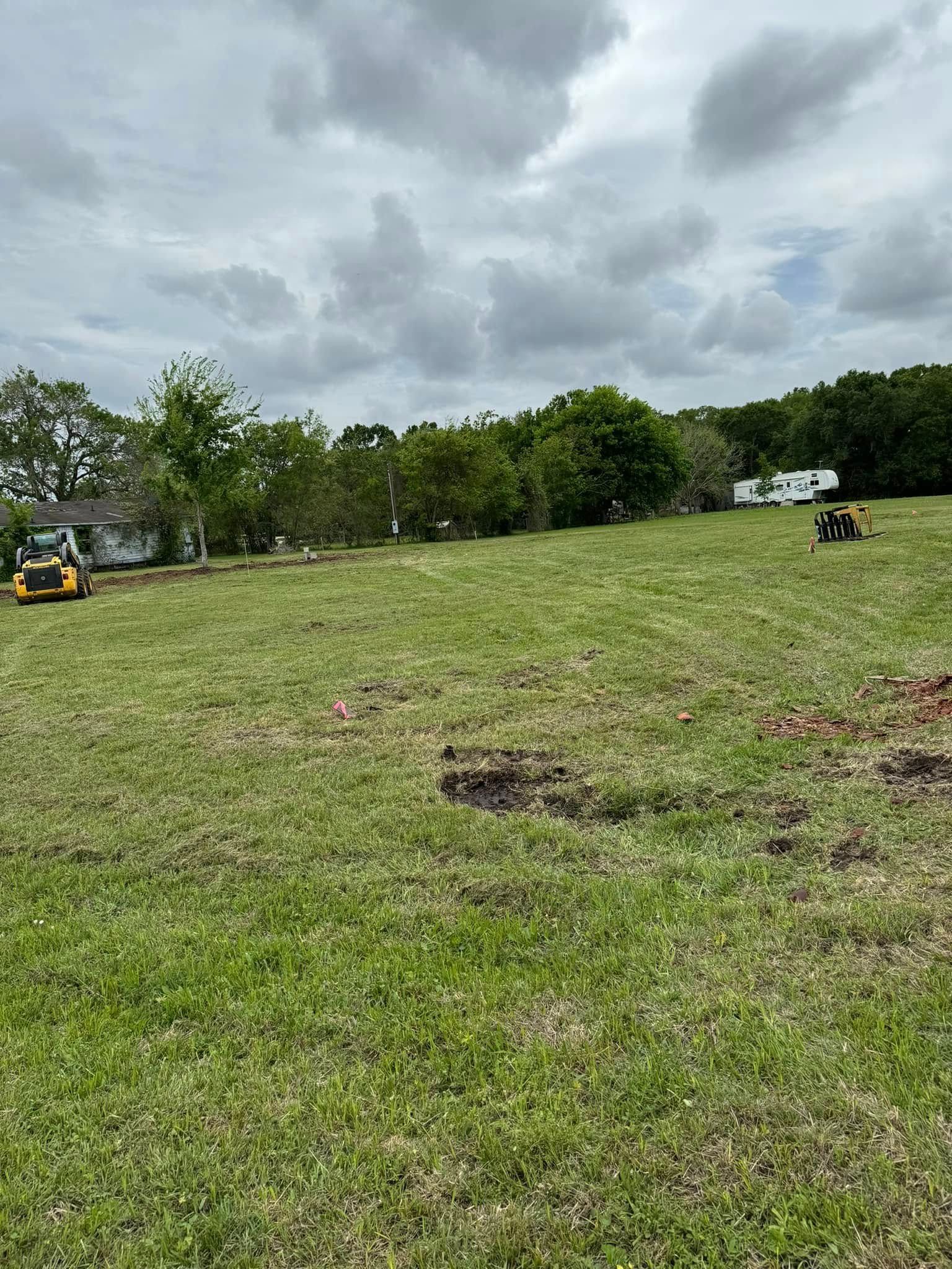Vast grassy field with two yellow lawn mowers; trees and a cloudy sky in the background.