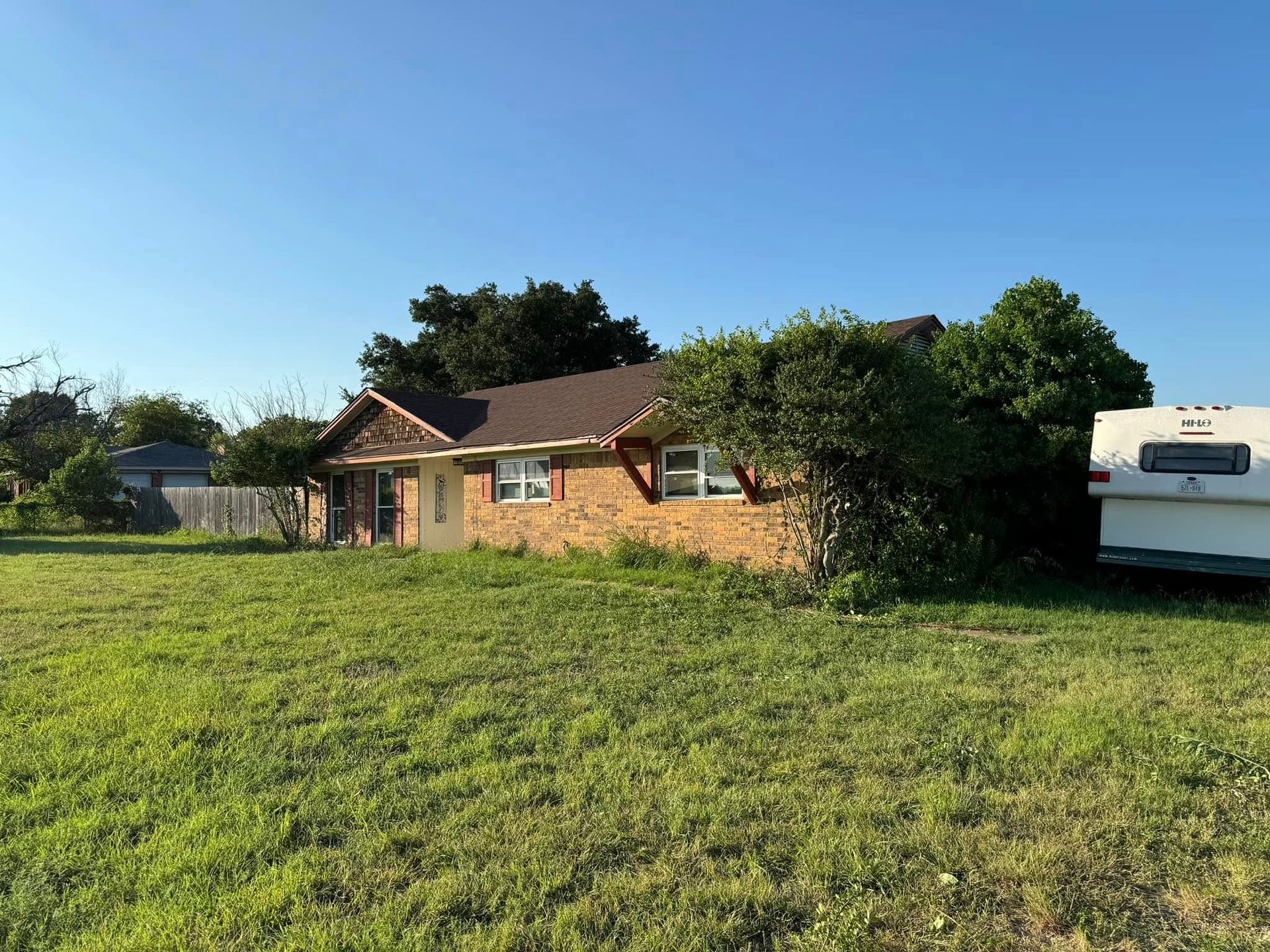 Single-story brick house with brown roof and overgrown yard under a blue sky. A camper is parked to the right.