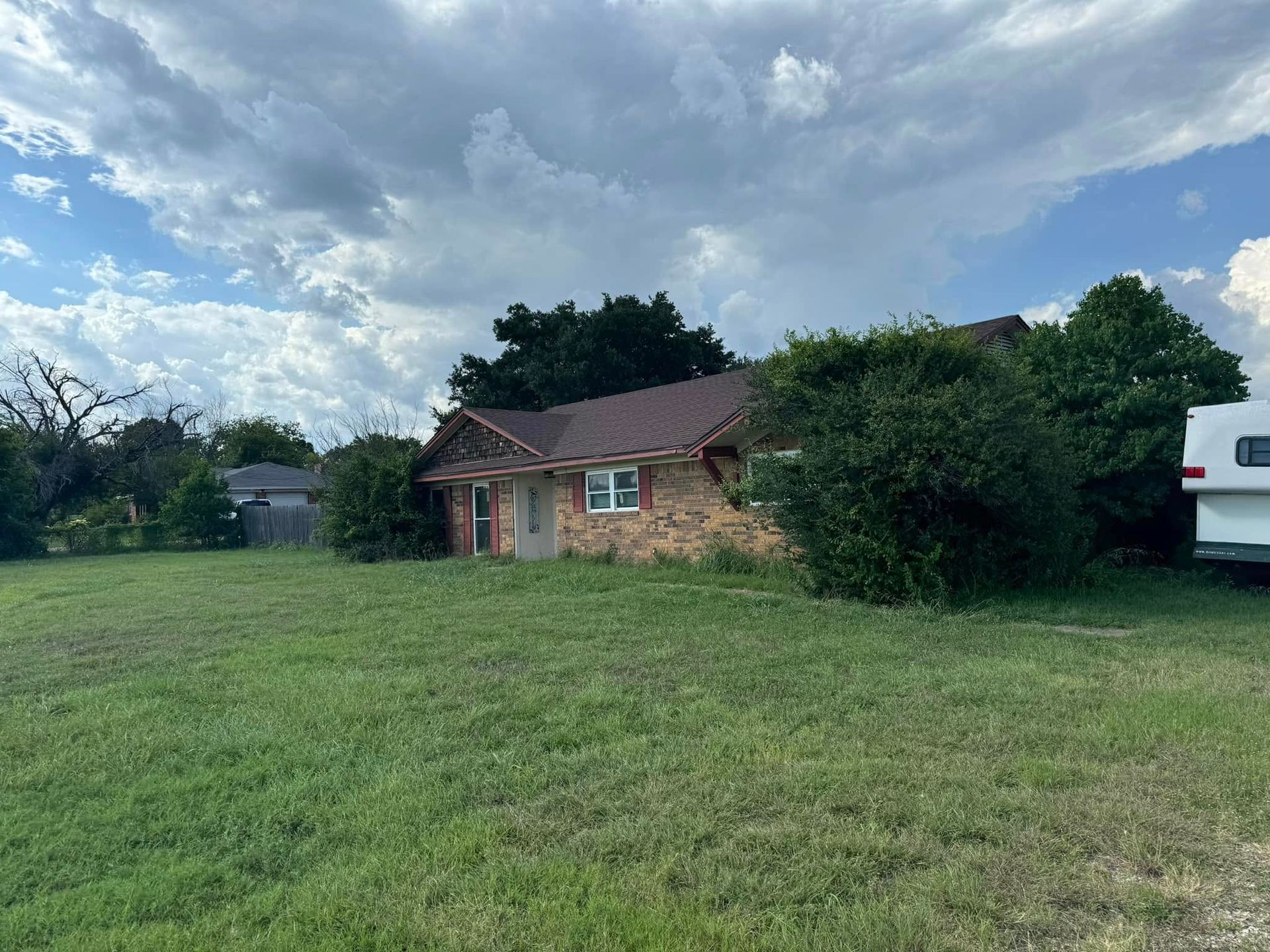 A brick house with a brown roof and overgrown shrubs under a cloudy sky.