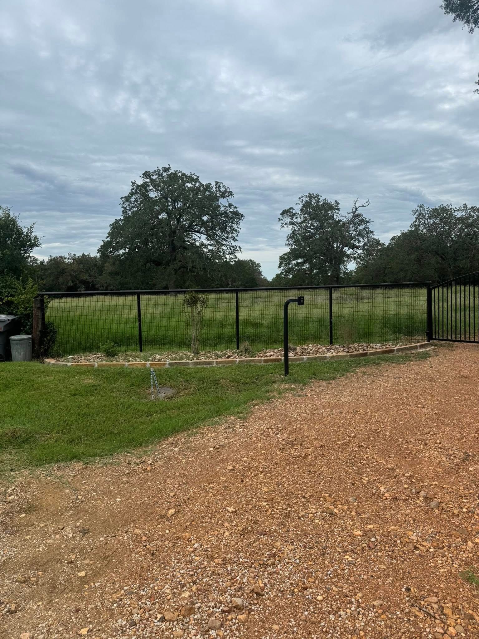 Black fence in front of a green field, with trees in the background under a cloudy sky.