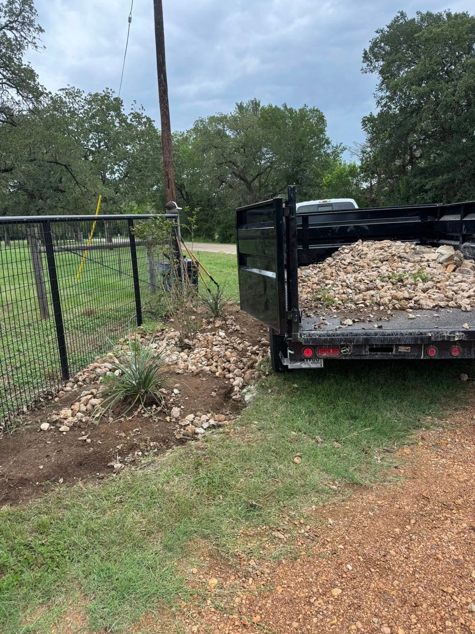 Dump trailer filled with yard waste beside a black chain-link fence on a cloudy day.