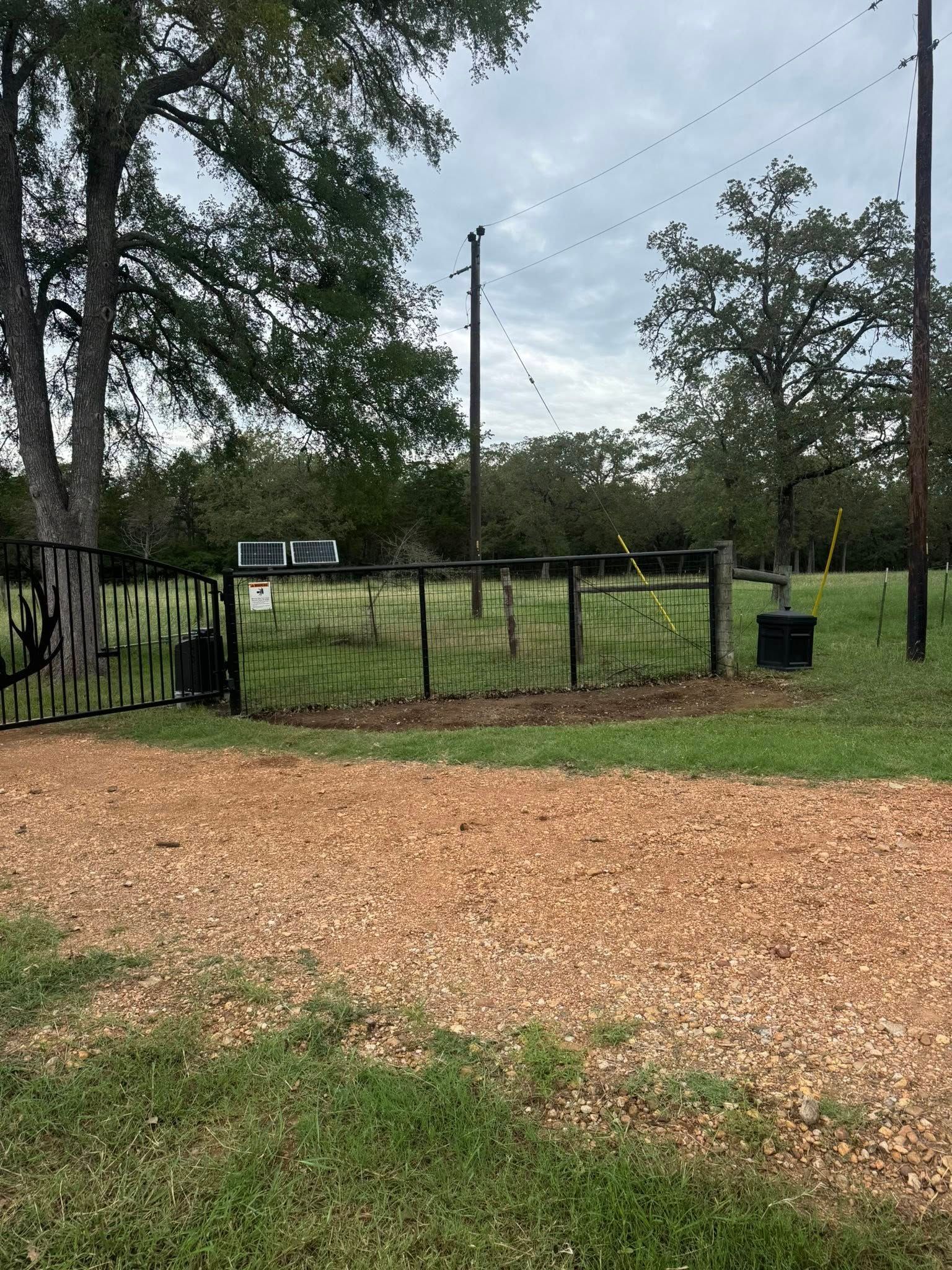 Gravel driveway leading to a gate, open, with a fenced pasture and a distant building under a cloudy sky.