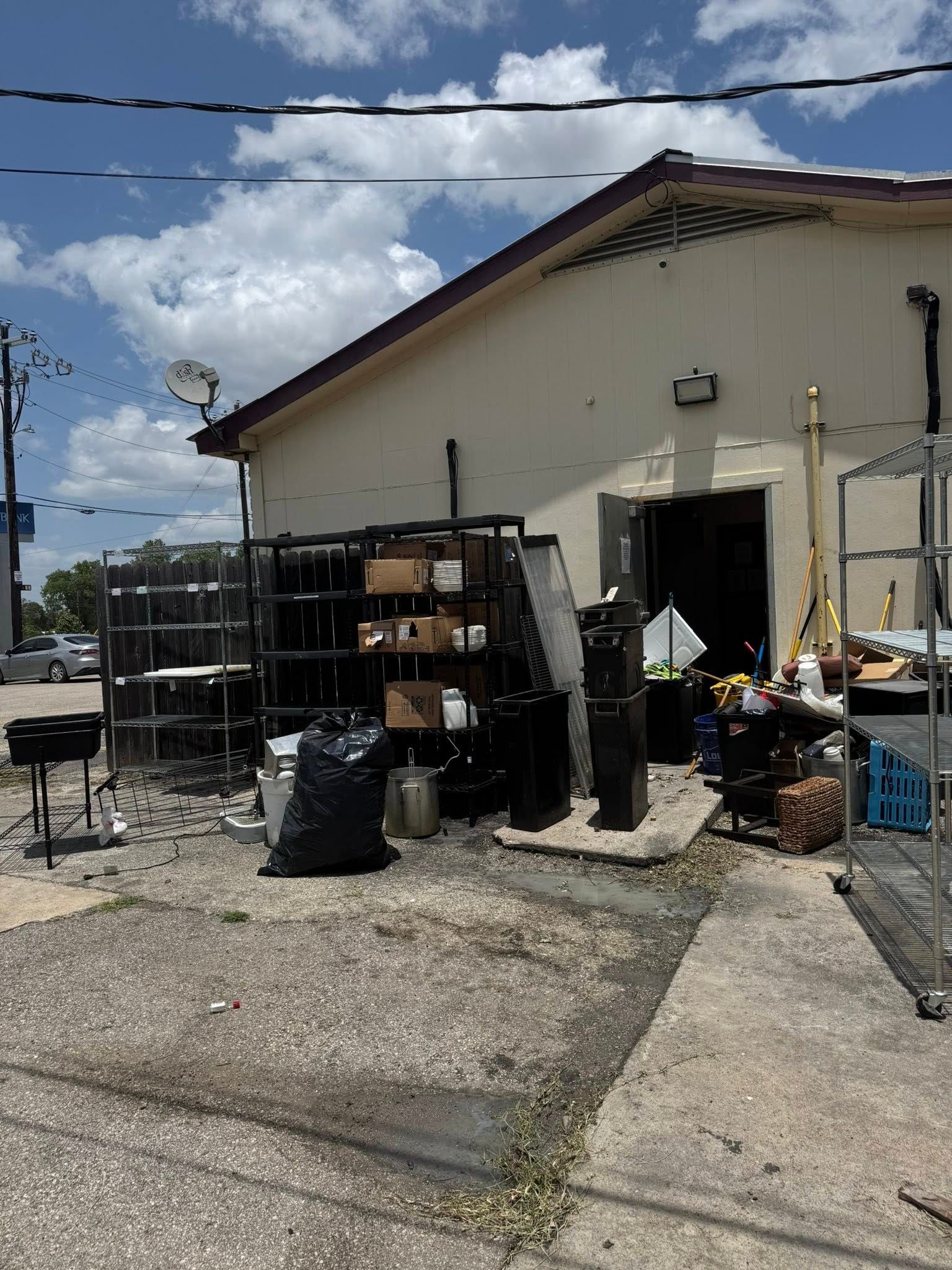 Exterior of a building with cluttered outdoor storage; shelves, boxes, black trash bag, and various items. Bright, sunny day.