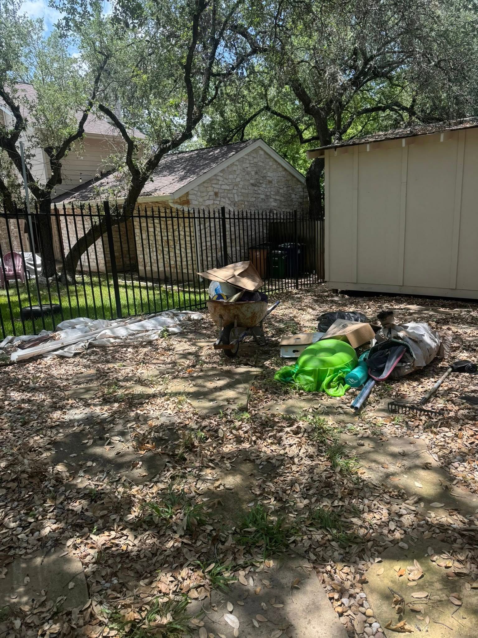 Backyard with debris, including cardboard boxes and plastic bags, near a wooden shed and black fence.