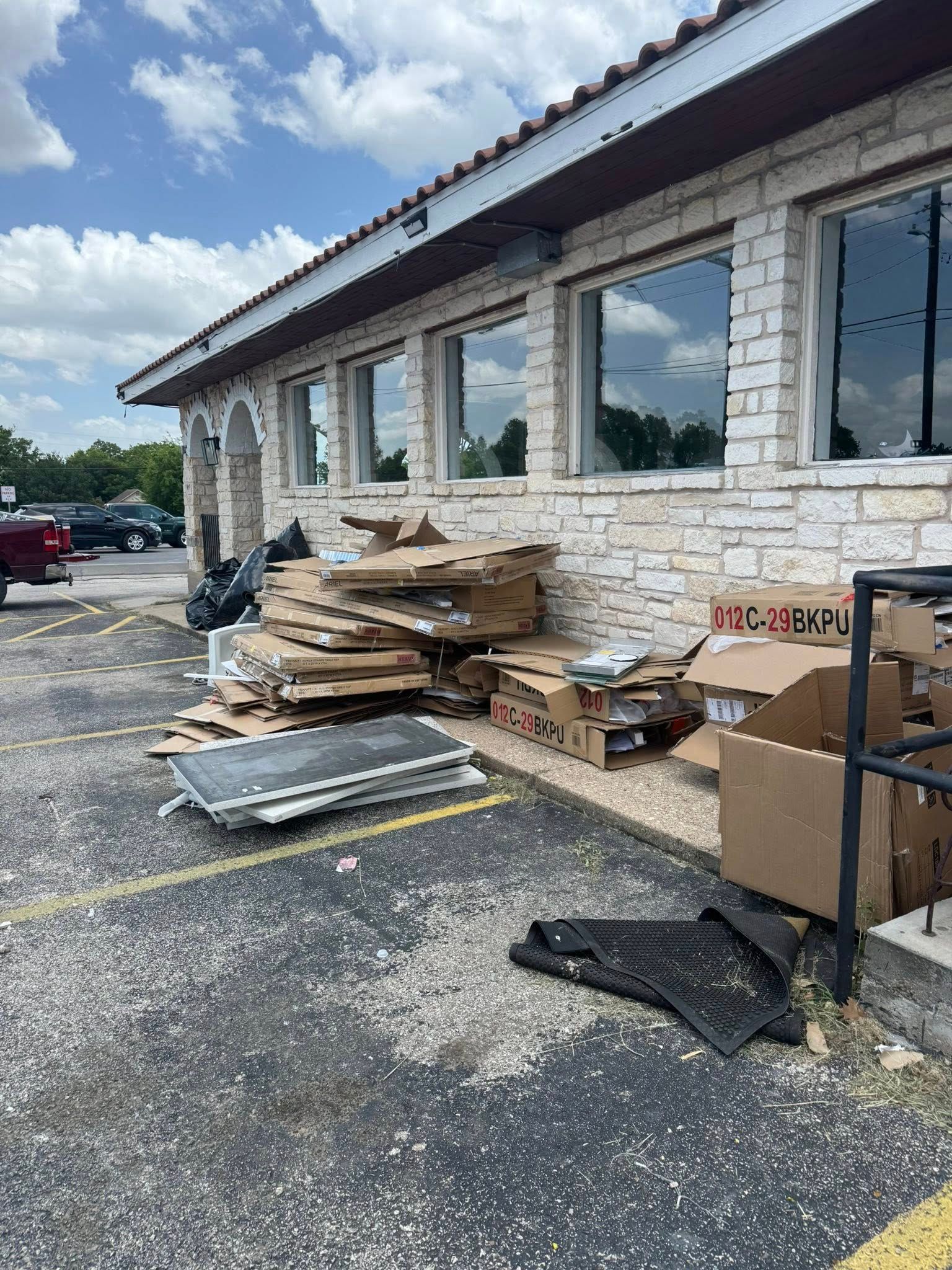 Cardboard boxes and debris piled outside a building with stone siding, on a paved parking lot.