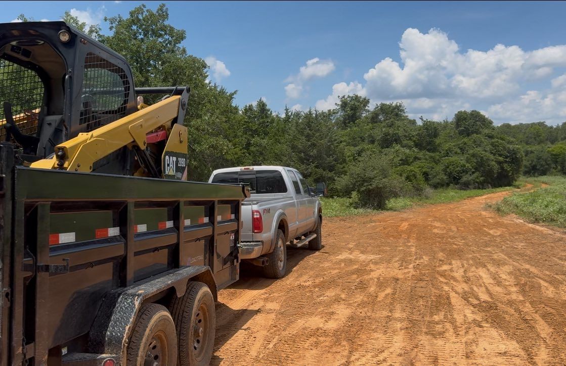 A truck pulling a trailer with a yellow skid steer on a dirt road, trees in the background, sunny day.