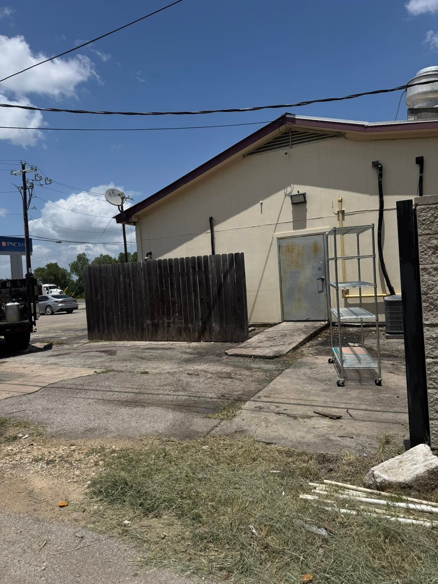Back alley with a tan building, wooden fence, and metal cart on a concrete surface. Blue sky overhead.