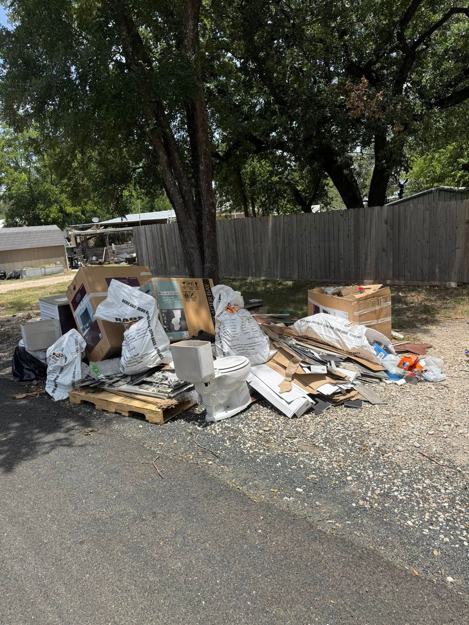 Pile of trash, including boxes and construction debris, sits near a tree and fence on a gravel surface.