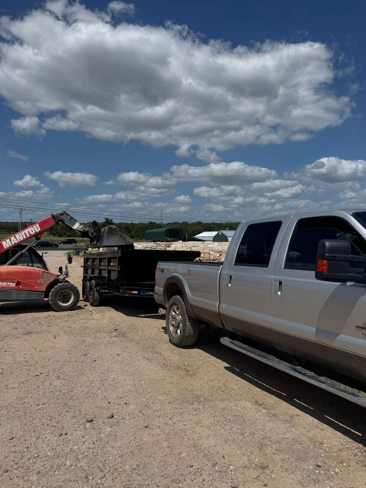 Silver pickup truck towing a trailer with a forklift in the background on a gravel surface under a blue sky with clouds.