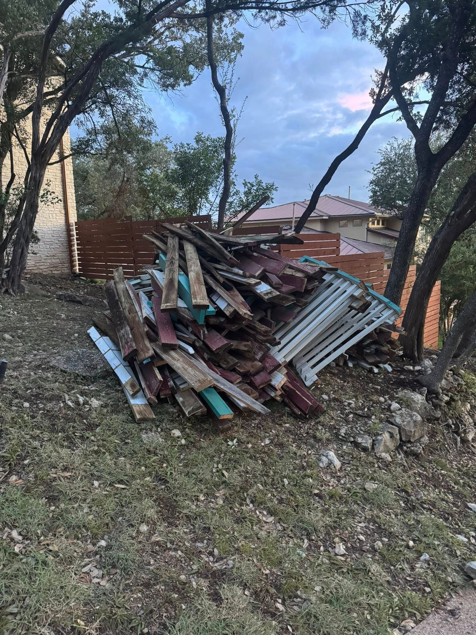Pile of wood and metal debris on a grassy hillside, trees in background, houses in the distance.