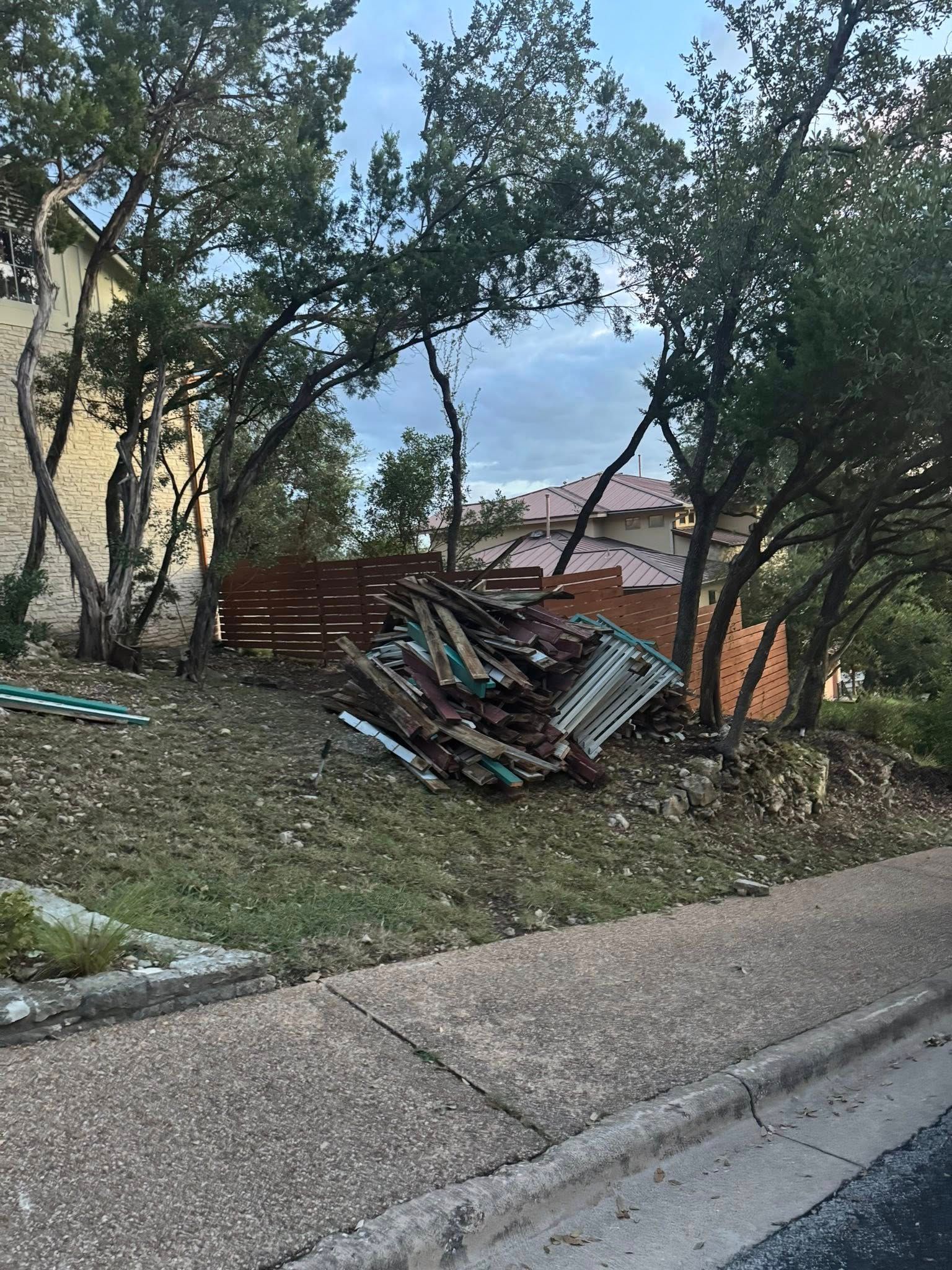 Pile of damaged, corrugated metal sheets on a grassy hillside next to a sidewalk and trees. Buildings visible in the background.