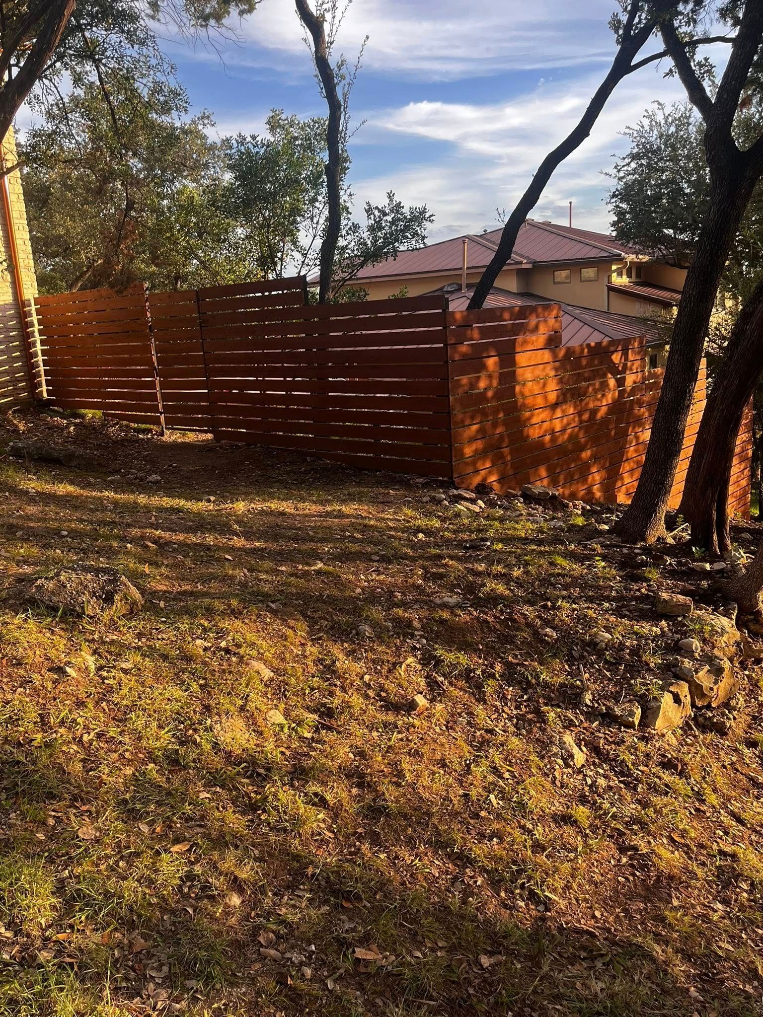 Wooden fence in a yard with trees, autumn foliage, and a house in the background on a sunny day.