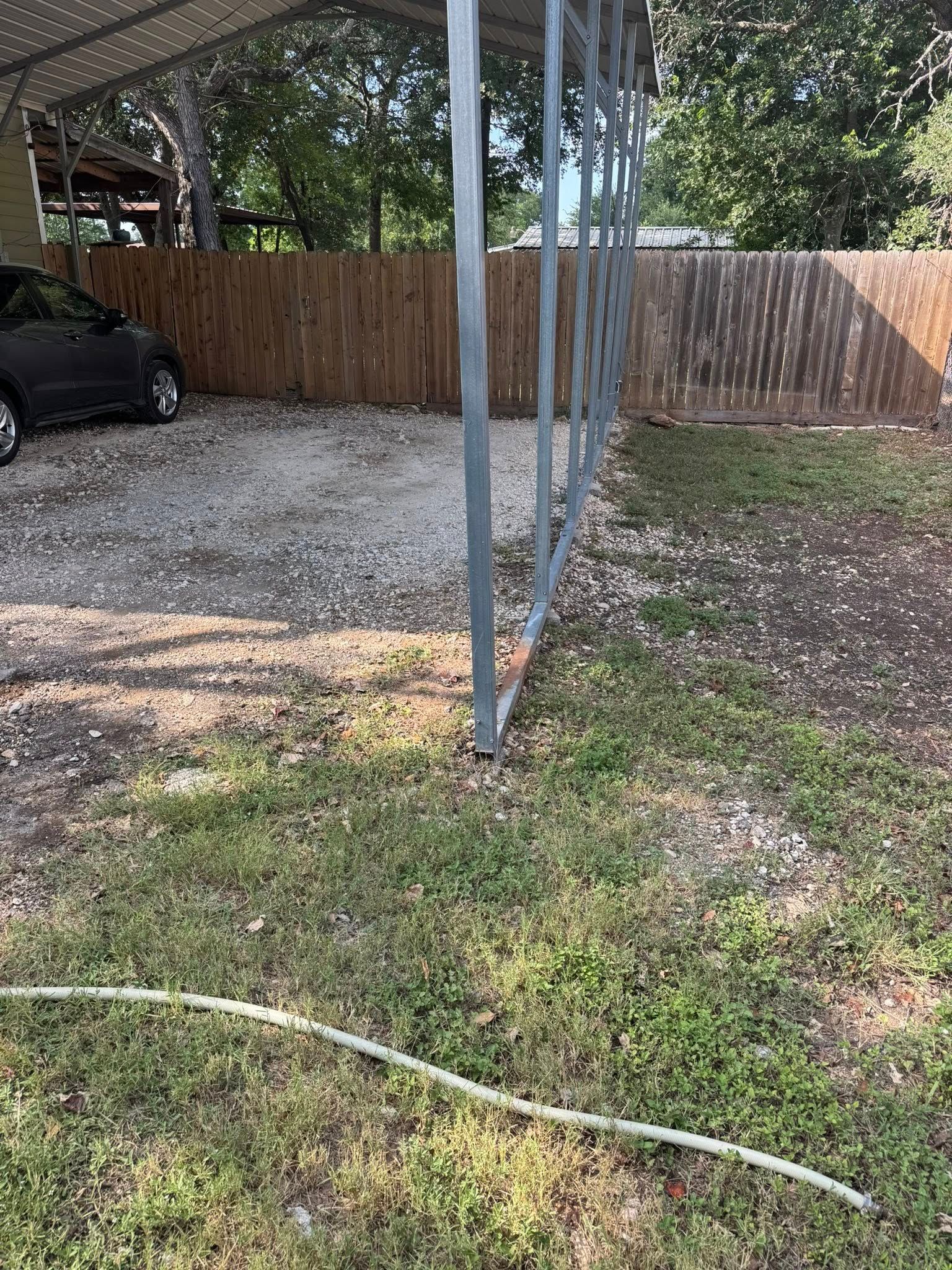 Carport with a parked car and gravel driveway, beside a wooden fence and grass lawn.