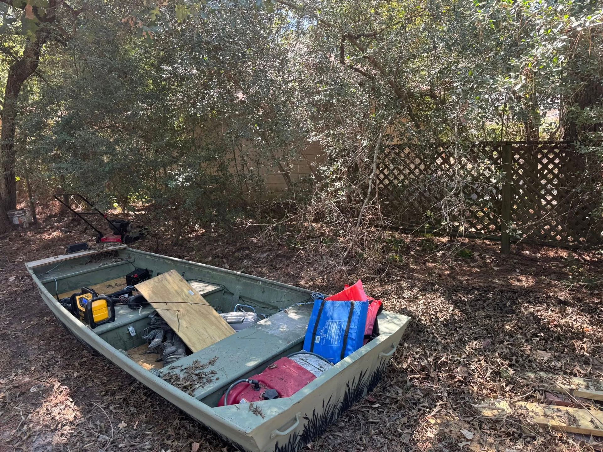Green boat on dry land, with debris inside. Wooden fence and trees in the background.