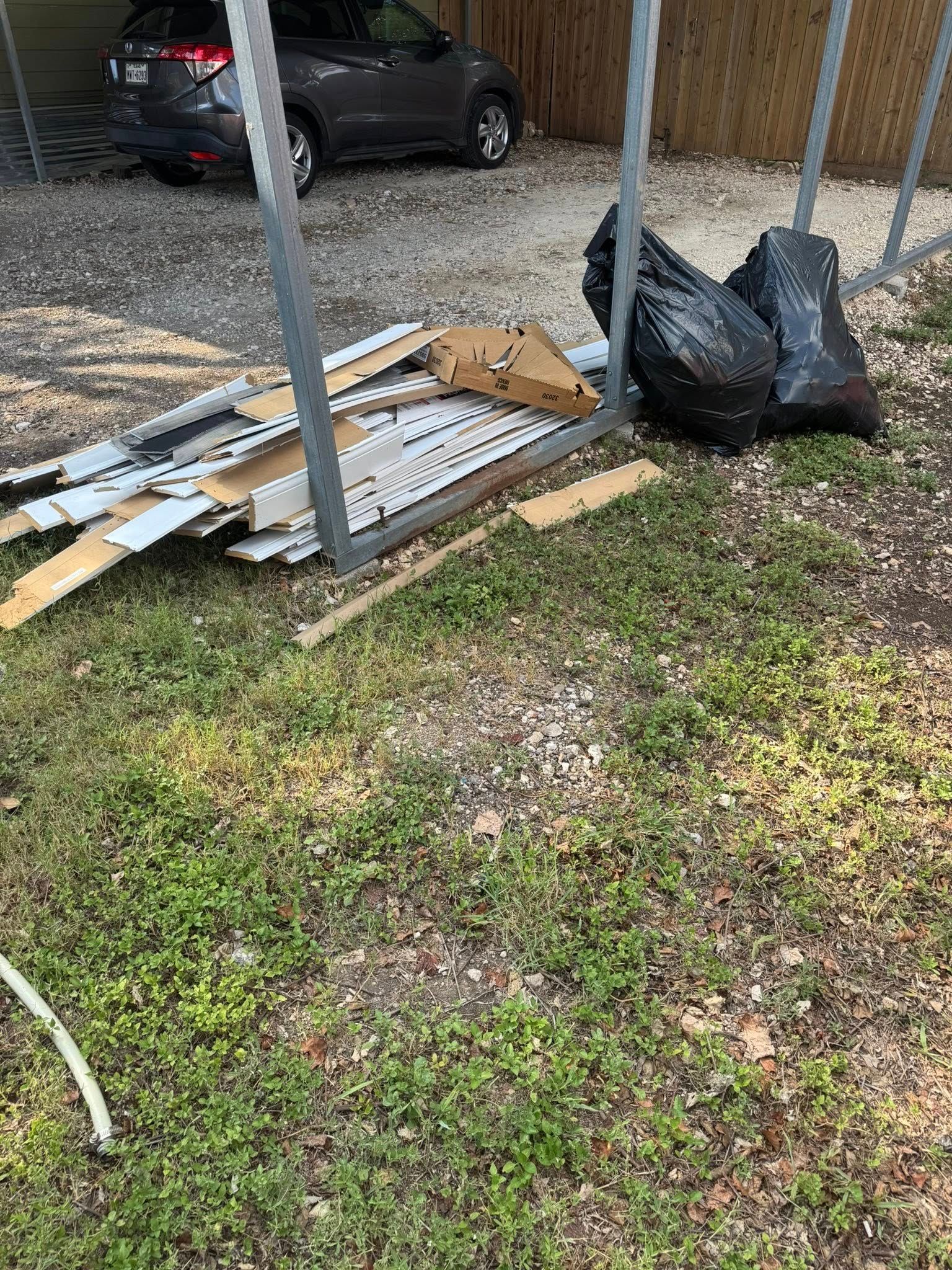 Pile of debris, including wood and cardboard, next to black trash bags and a gray car under a carport.