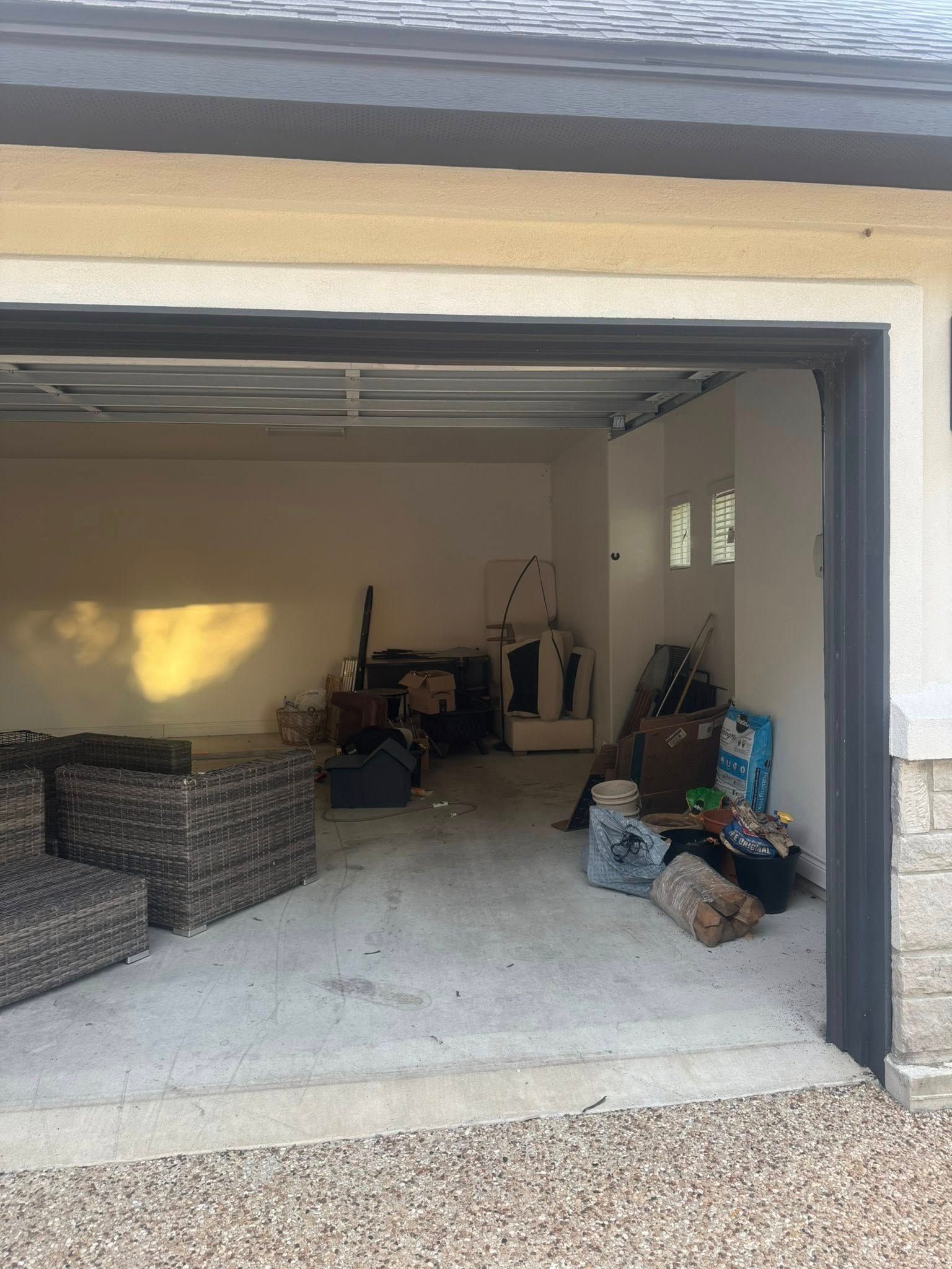 Open garage cluttered with furniture, bags, and boxes. Beige walls, concrete floor, and grey trim.