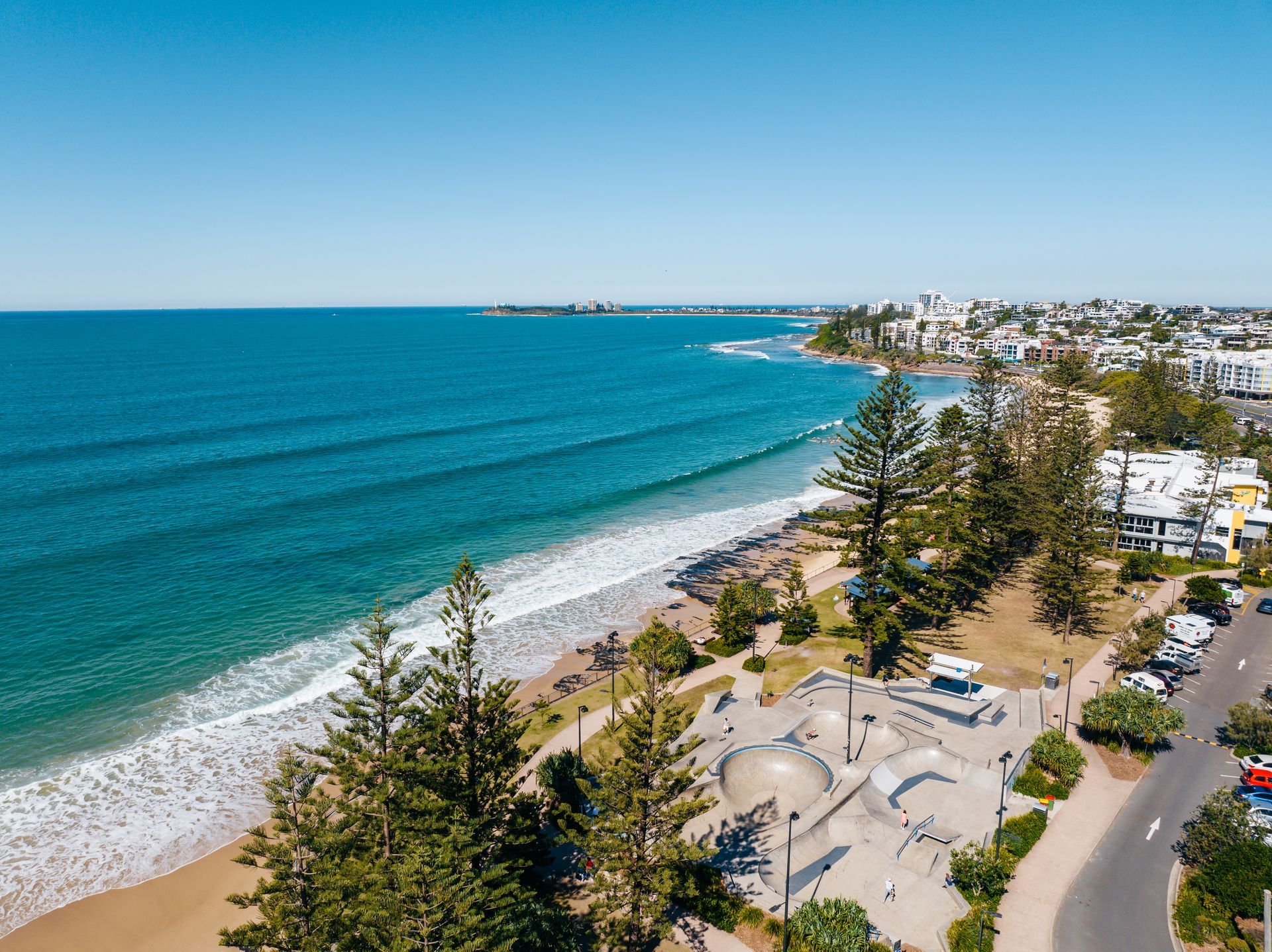 Beachfront aerial view with ocean waves, skate park, and coastal town under a blue sky.
