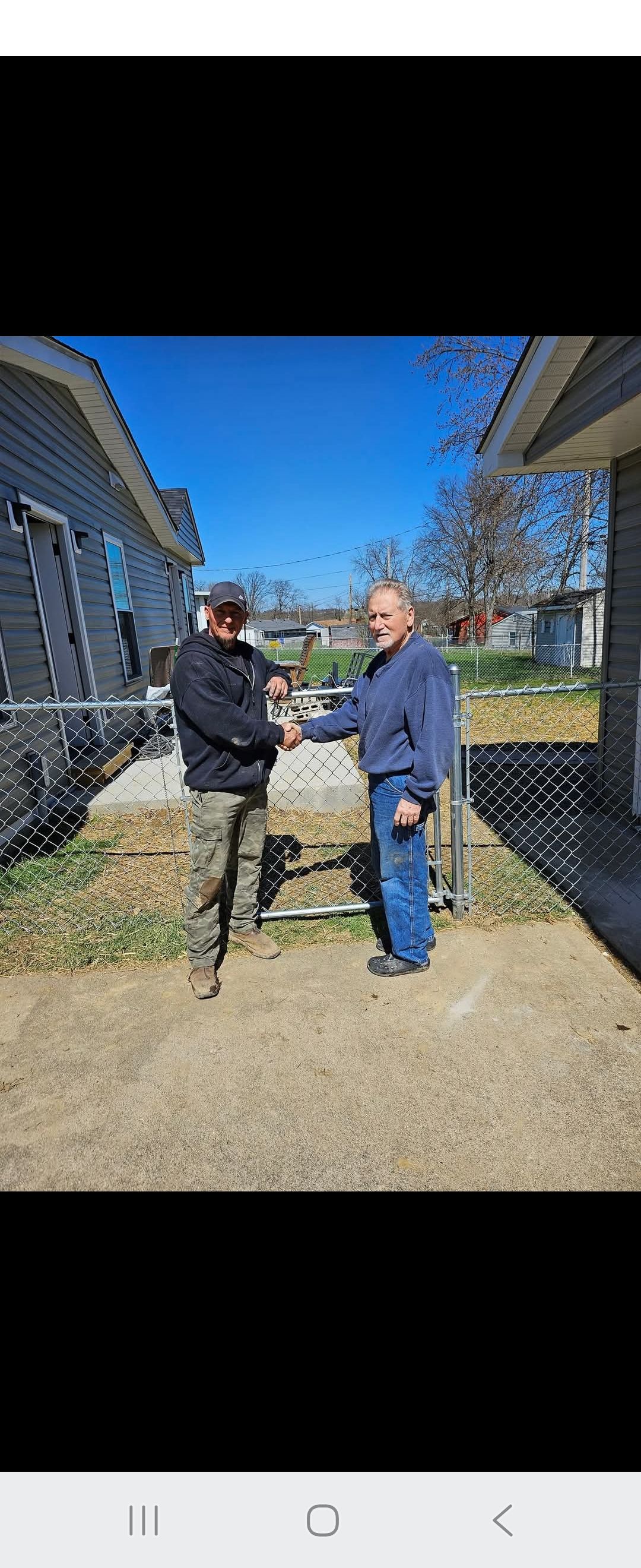 Two men shaking hands in front of a chain-link fence on a sunny day.