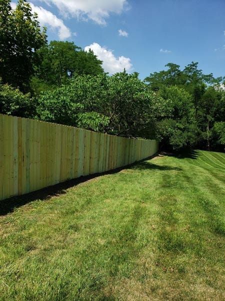 Wooden fence along a grassy slope, with trees in the background under a blue sky.