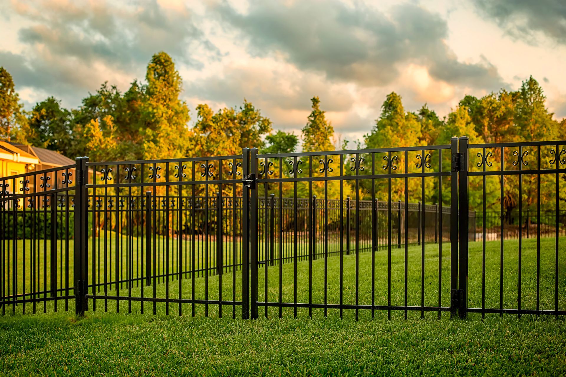 Black metal fence in a green yard, with trees and a house in the background under a cloudy sky.