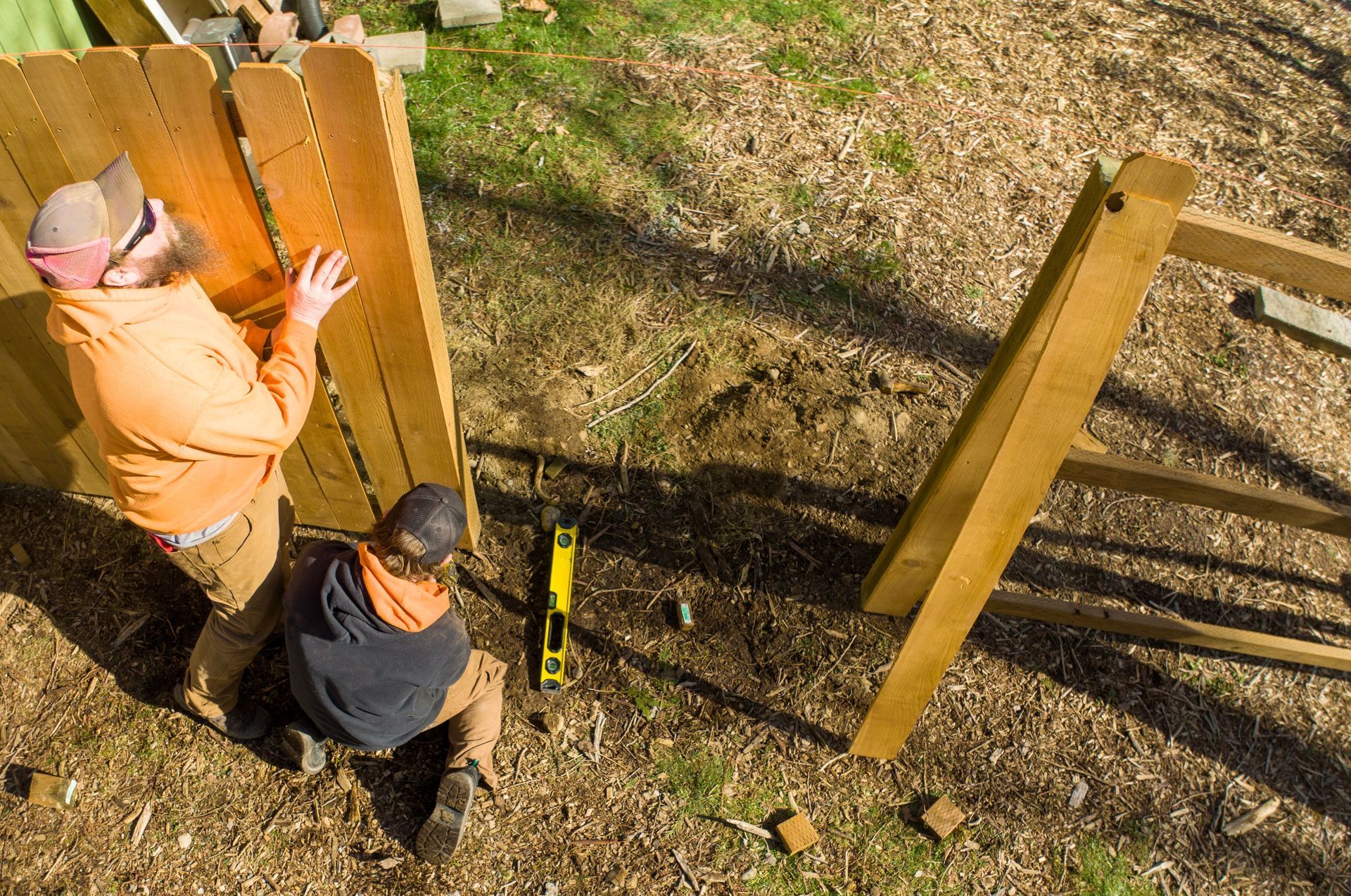 Two people building a wooden fence outdoors on wood chips; one person standing, one kneeling with a level.