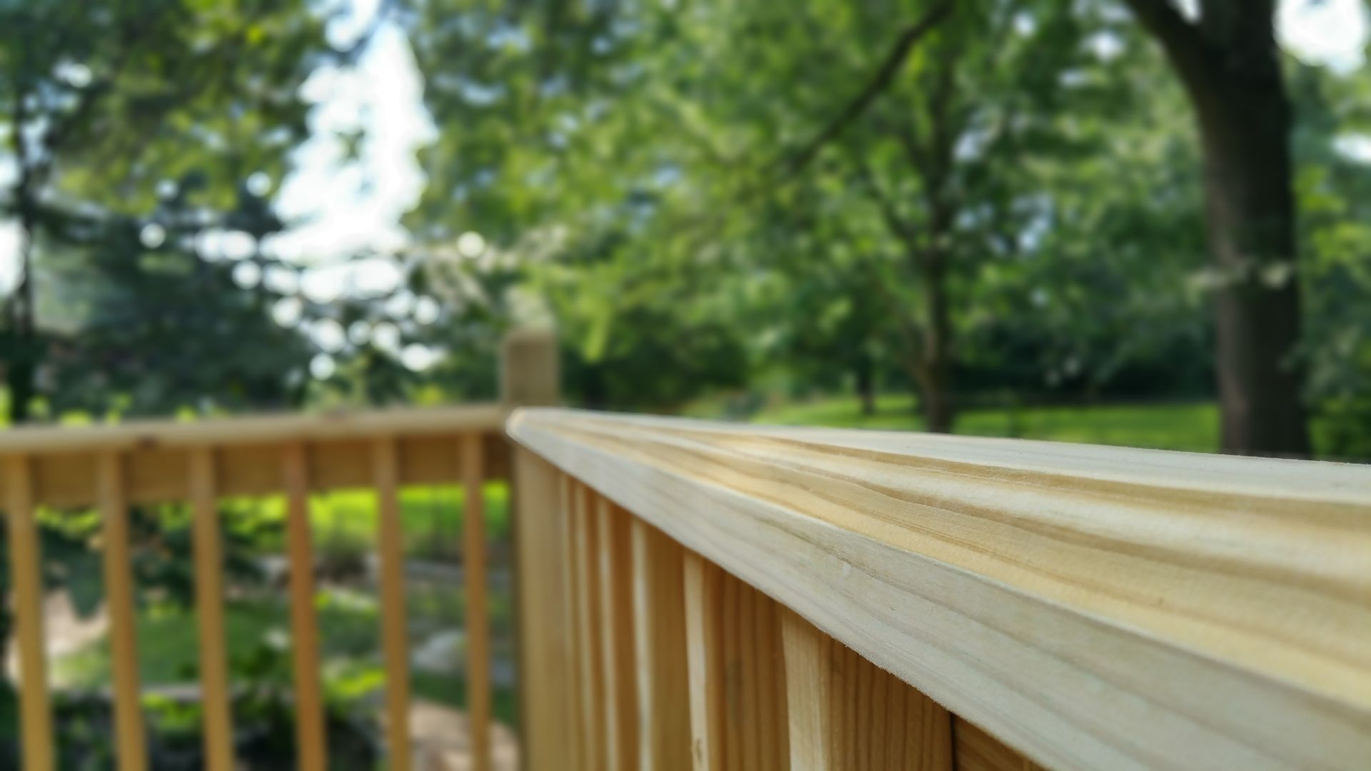 Wooden deck railing with a blurred background of green trees and grass.