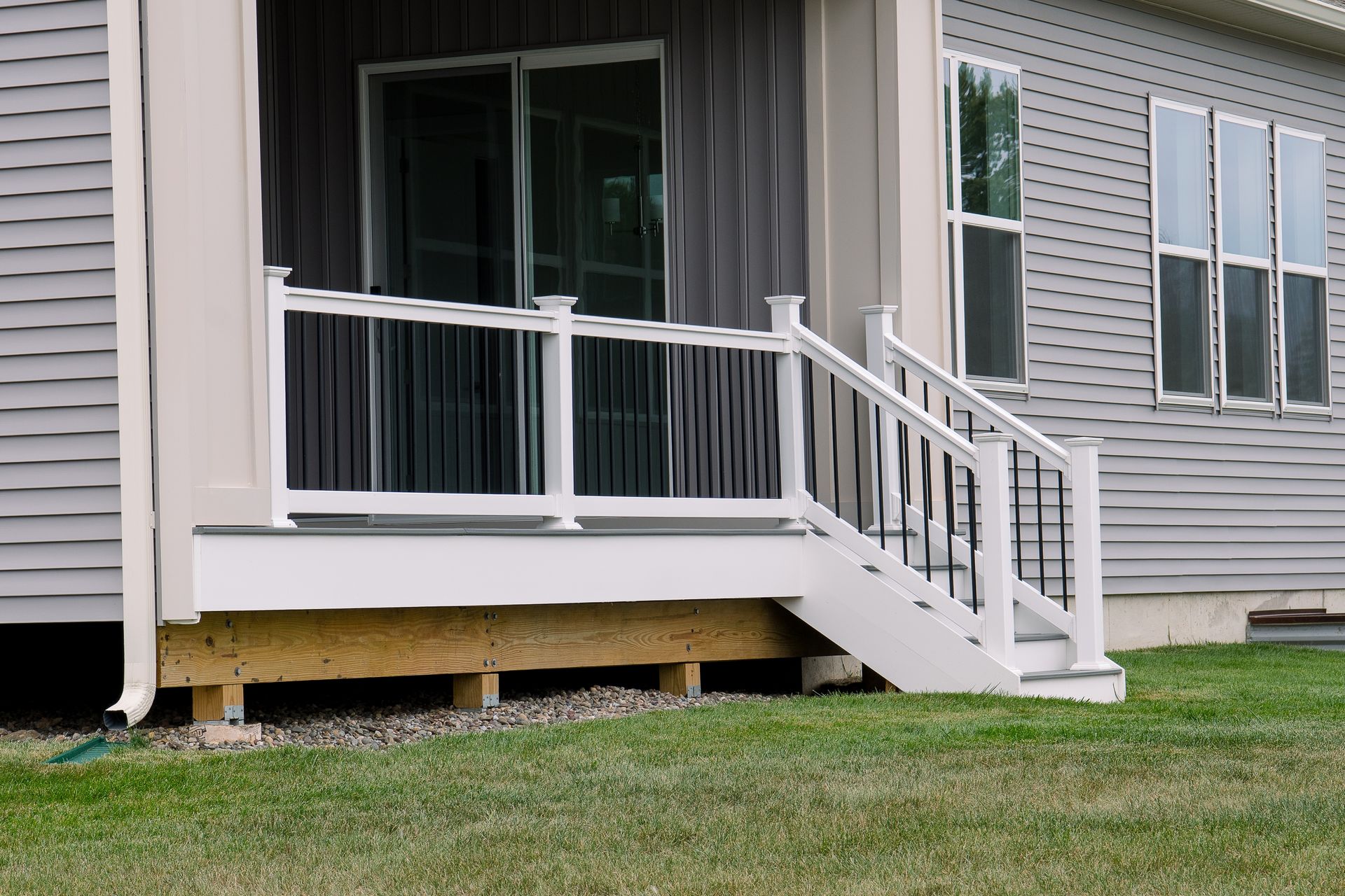 White porch with stairs and railing leading to a gray house with windows; green grass in the foreground.