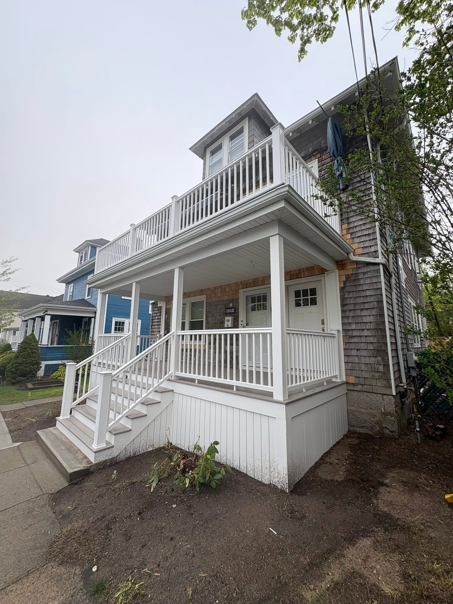 White two-story house with a front porch, balcony, and stairs beside a gravel yard