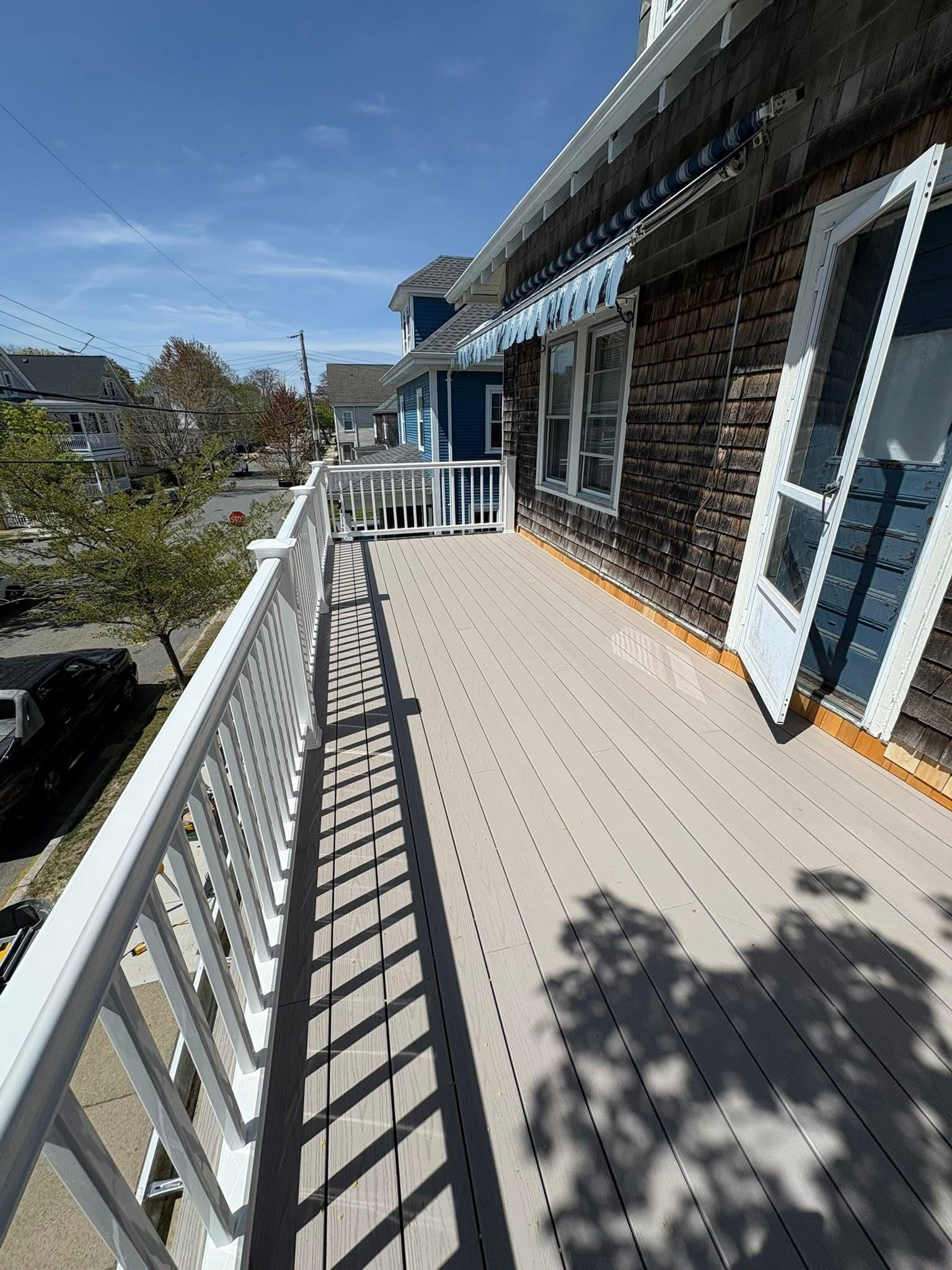Sunlit balcony with white railing, brick wall, and gray deck beside a blue door