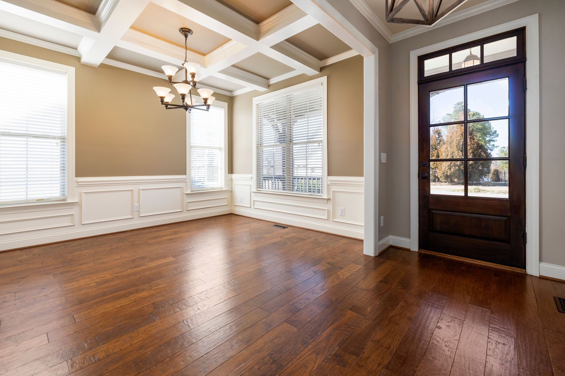 Empty dining room with hardwood floors, coffered ceiling, chandelier, and large windows beside a front door