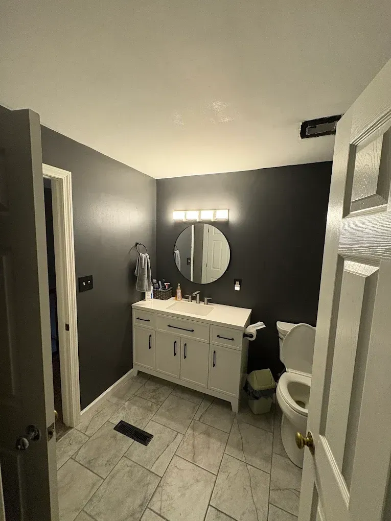 Modern bathroom with gray walls, white vanity, round mirror, lit vanity lights, and tiled floor.