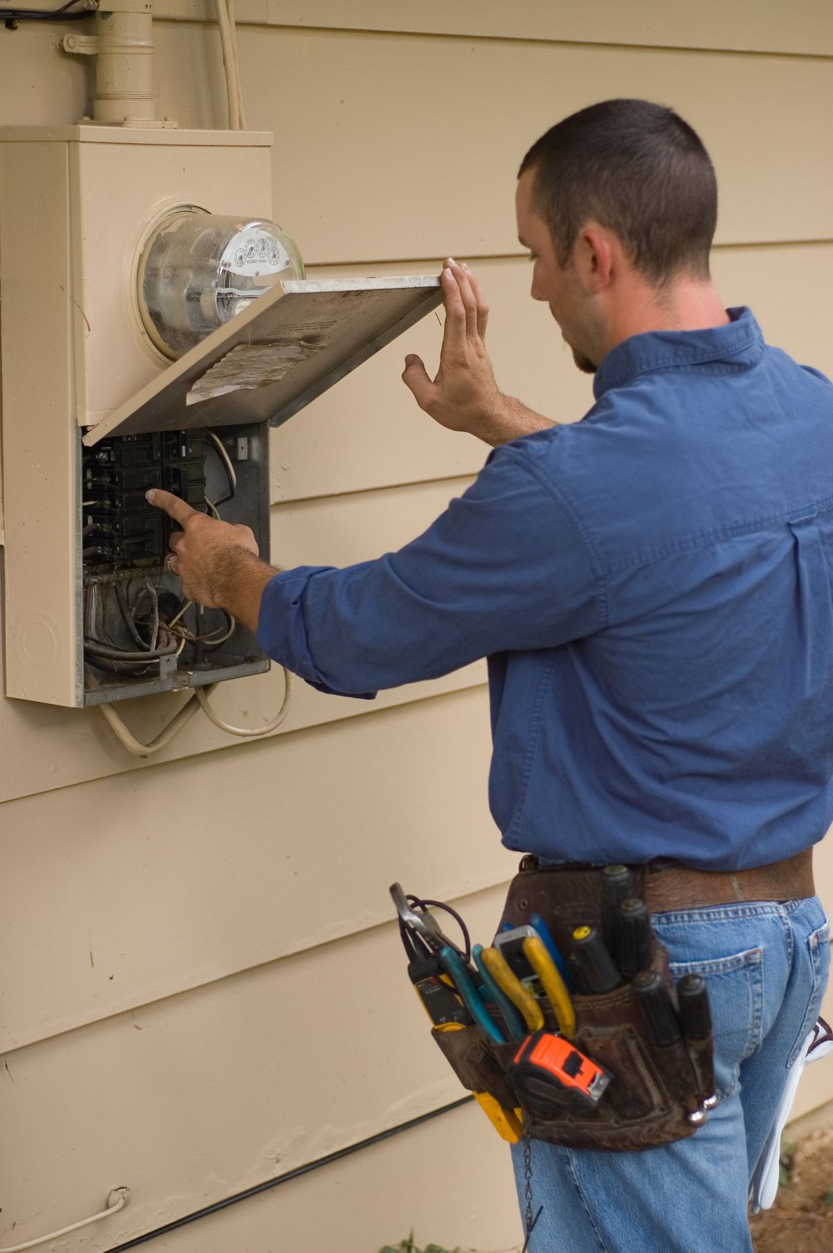 Electrician examining a circuit breaker panel on a building’s exterior. He's wearing a tool belt and blue shirt.