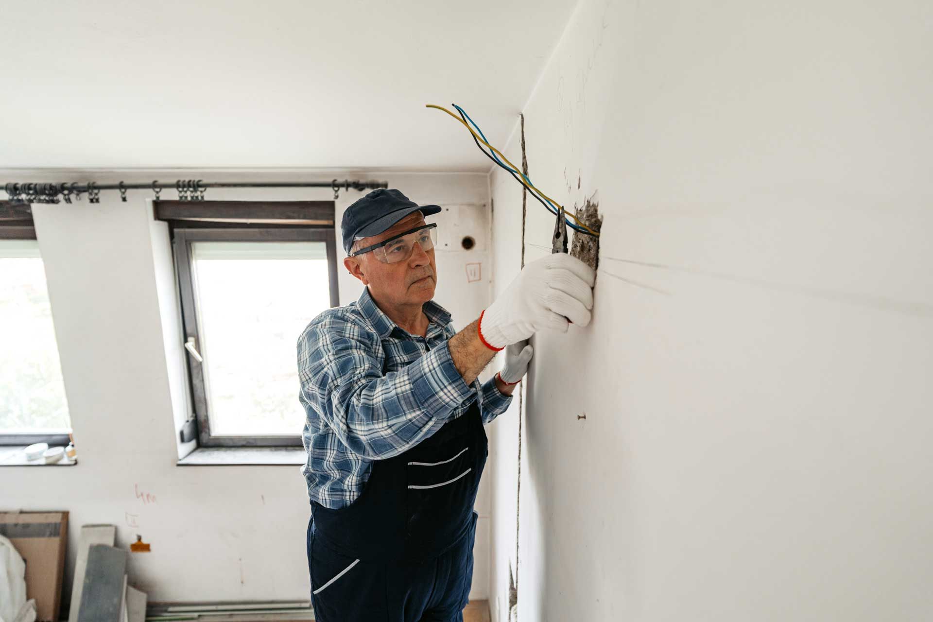 Electrician working on electrical wires inside a wall, wearing gloves and a cap.