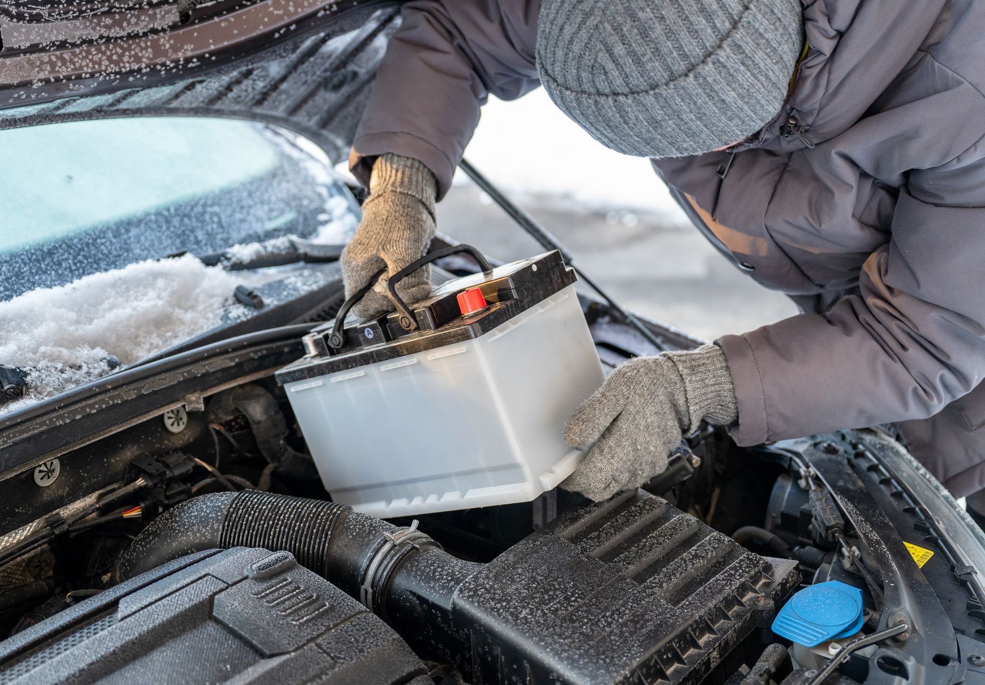 Person in winter gear working under a car hood with a plastic container and snow visible.