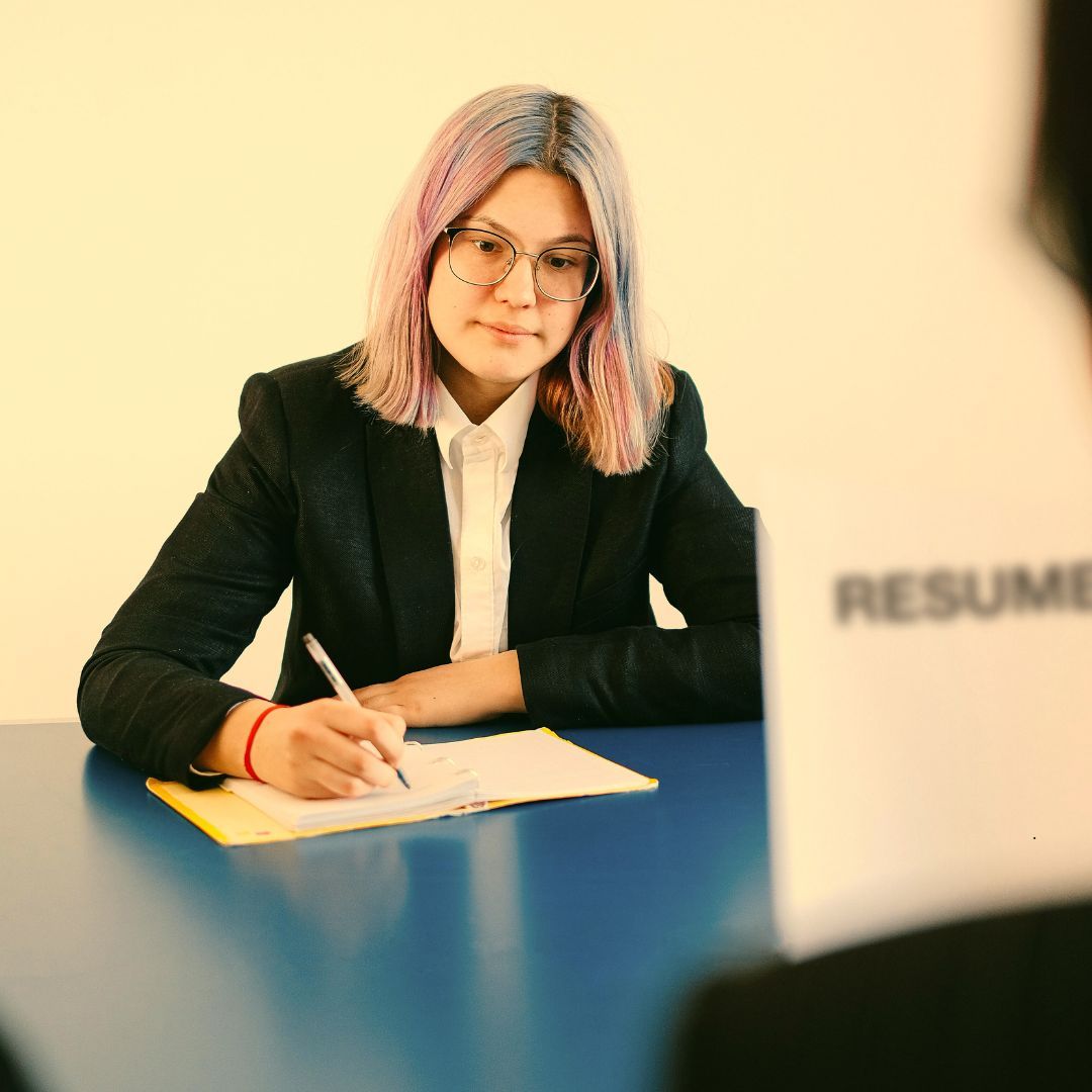 Woman in blazer writing at a desk, looking at a resume, focused.