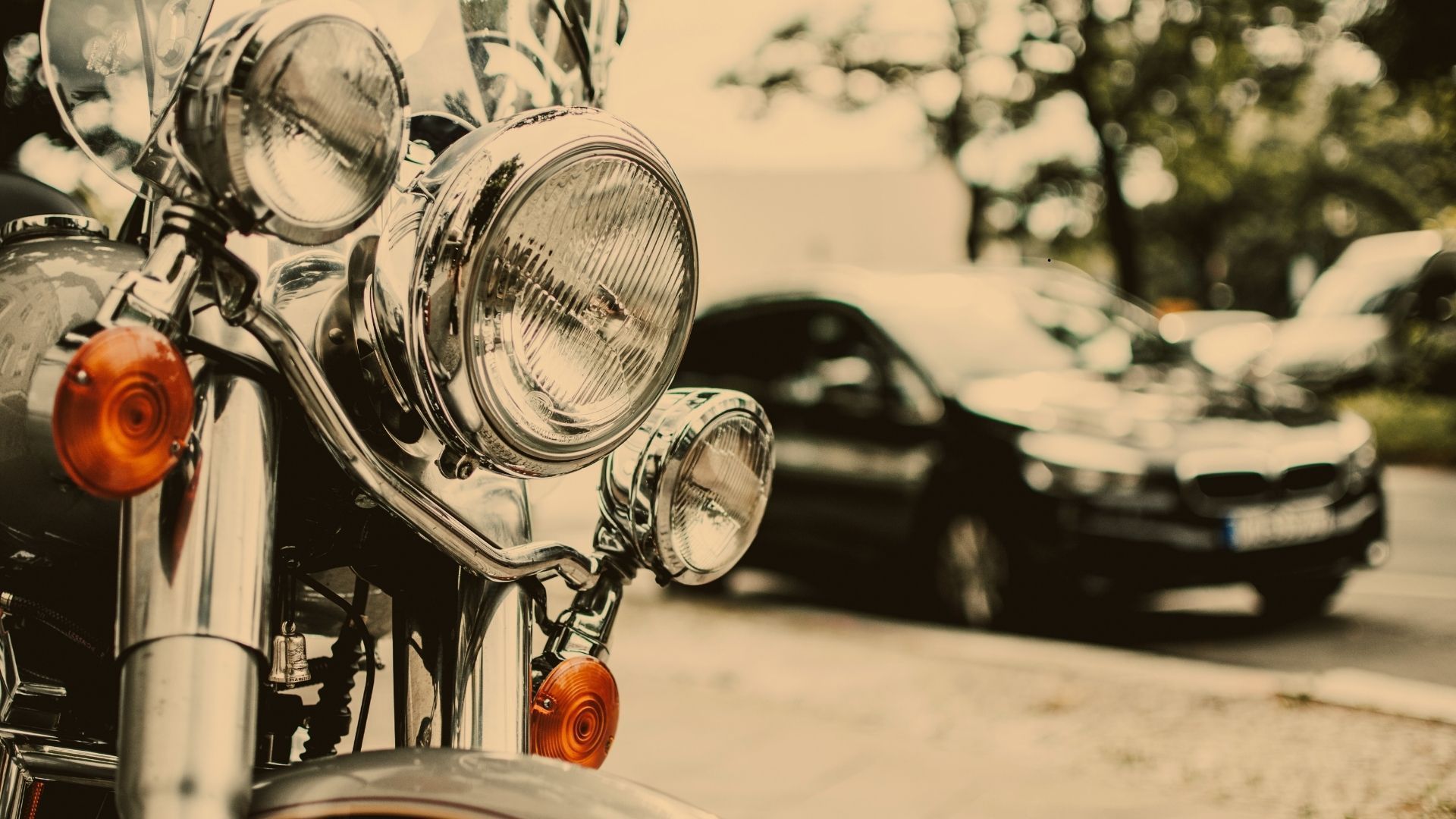 Chrome motorcycle headlights in foreground, black car blurred in the background on a city street.