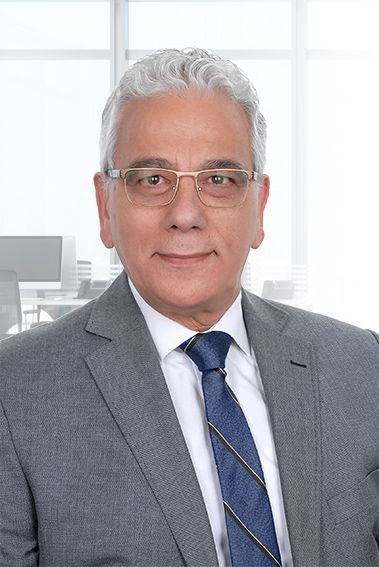 Man in gray suit, glasses, and blue tie smiles, with a bright office backdrop.