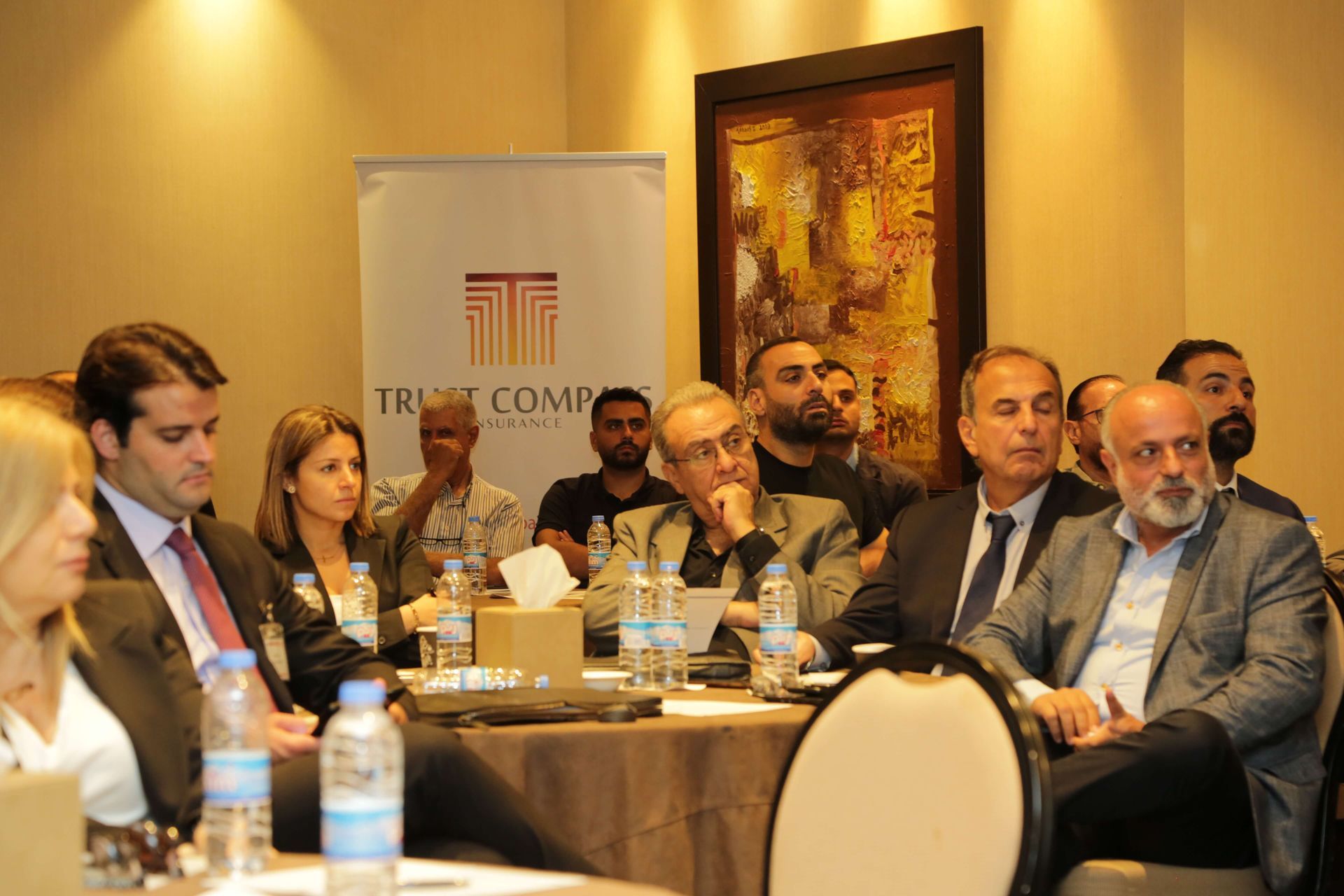 People seated around a table, attending a conference. Some are looking forward, others are talking. A banner is in the background.