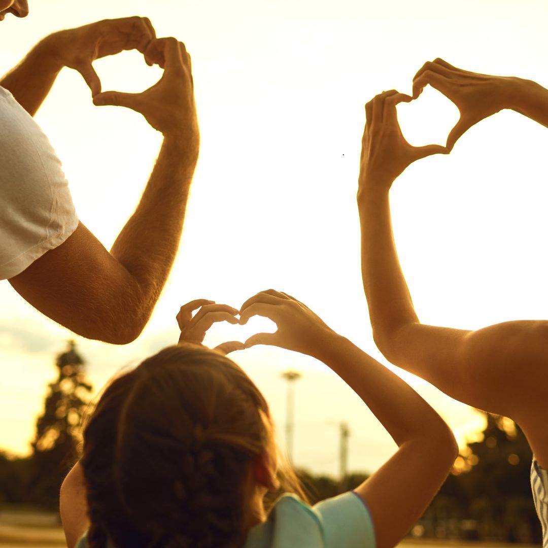 Family making heart shapes with their hands against a bright sky, possibly sunset.