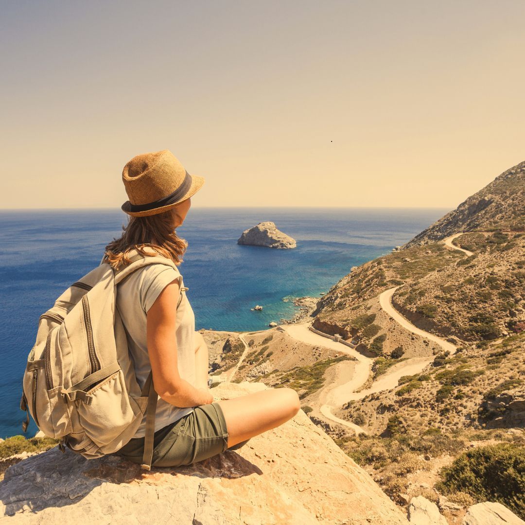 Woman in hat and backpack looks at ocean and winding road.