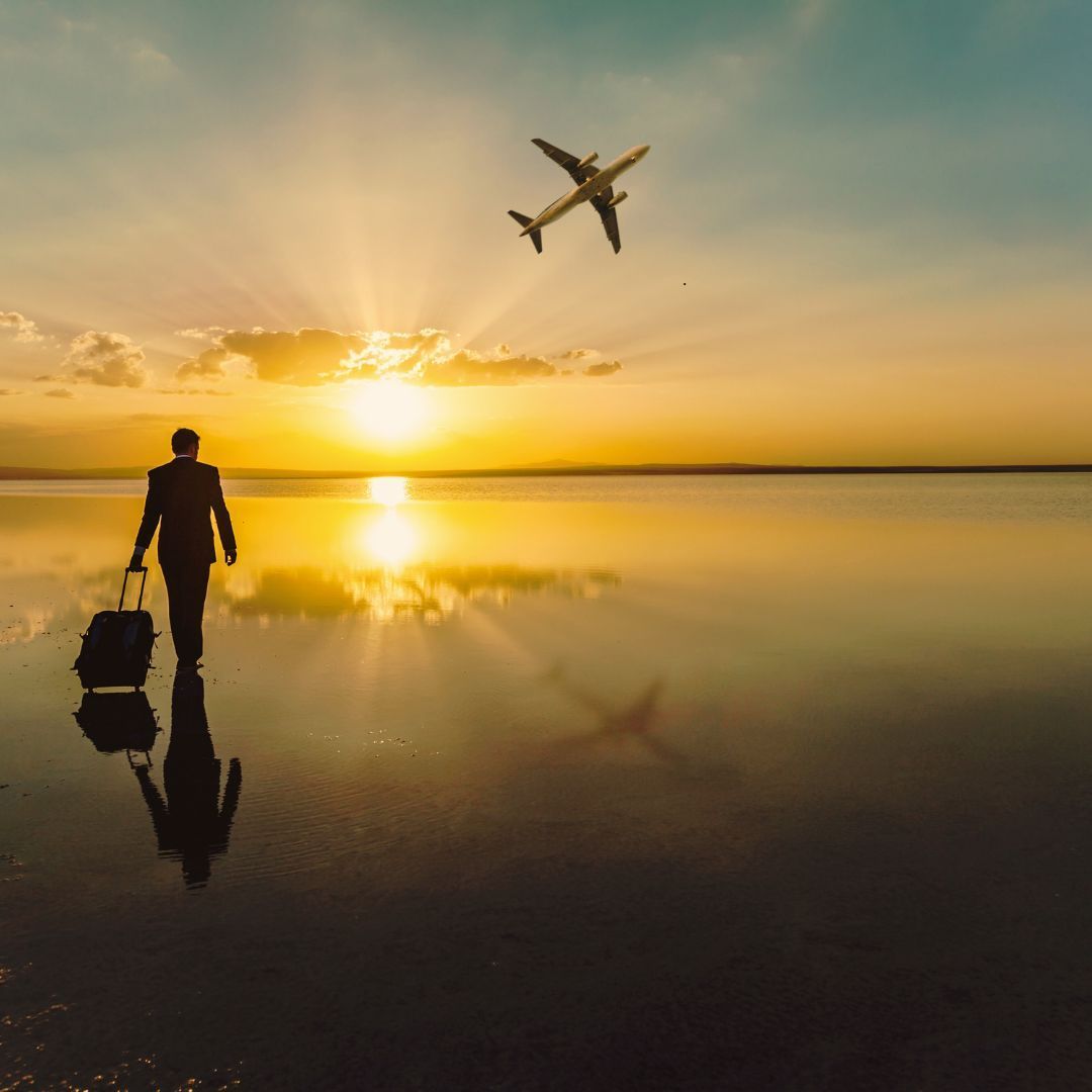 Silhouette of person with suitcase walking on water towards setting sun with airplane.