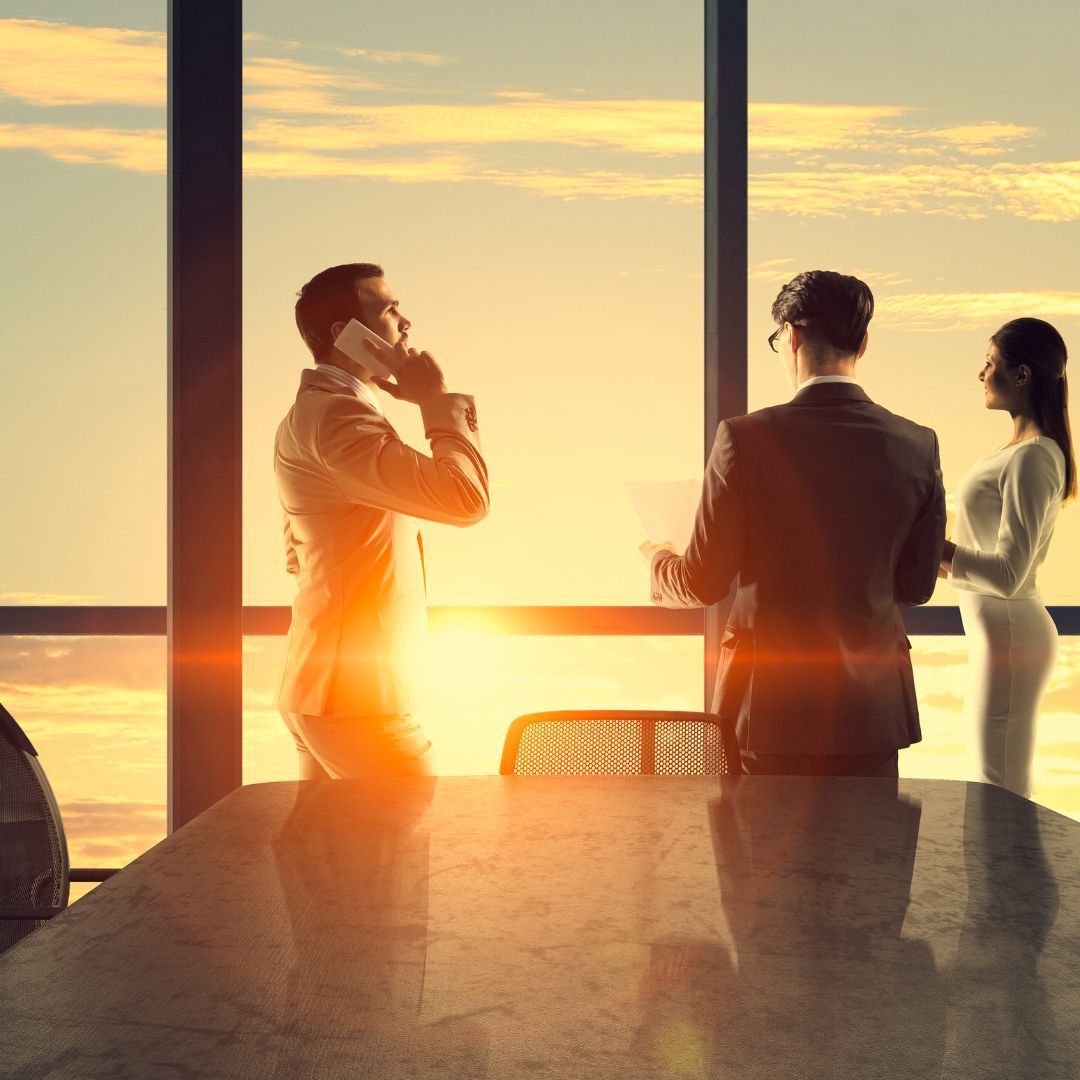 Businesspeople in suits at a conference table, overlooking sunset through a window; man on phone.