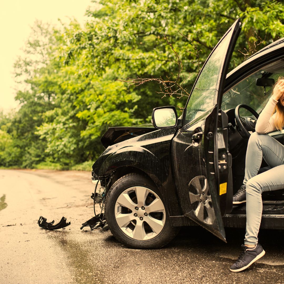 Woman in car after accident, damages visible. Black car on wet road, forest background. She looks distressed.
