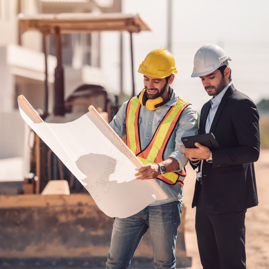 Two men at construction site, one in hard hat and vest, reviewing blueprints; other in suit using tablet.