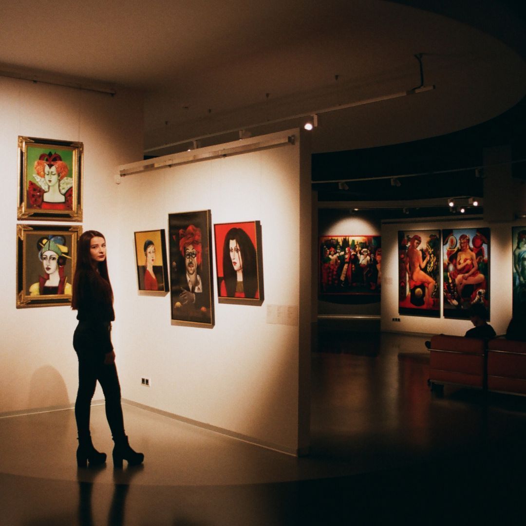 Woman in an art gallery, looking at paintings on the wall; warm lighting, dark shadows.
