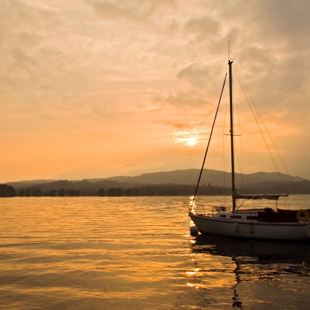 Sailboat on calm water at sunset, with golden light reflecting. Mountains in the distance.