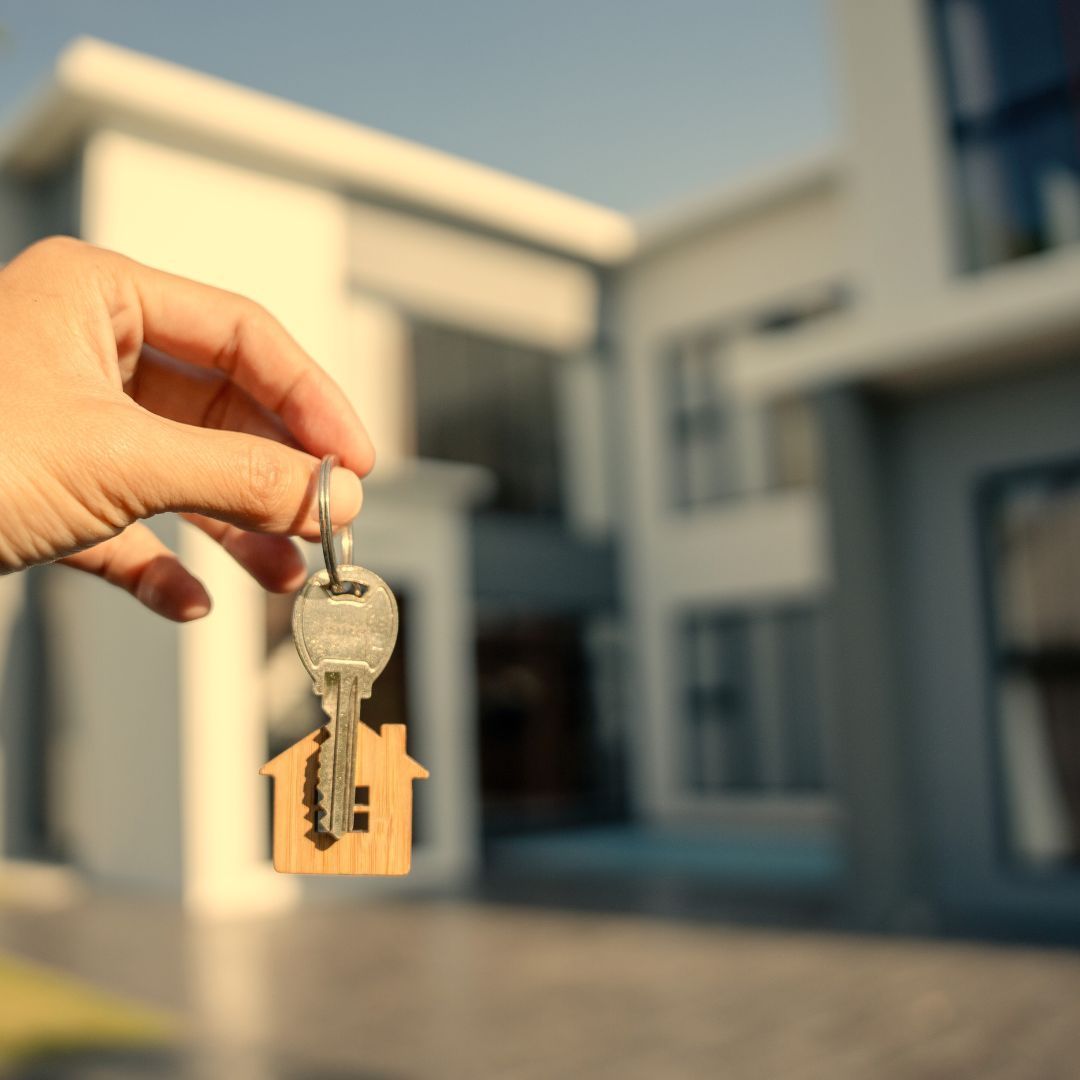Hand holding house keys in front of a modern home.