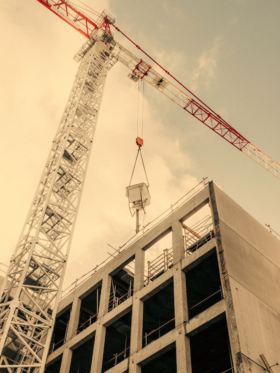 Construction crane lifting material to unfinished concrete building against a cloudy sky.