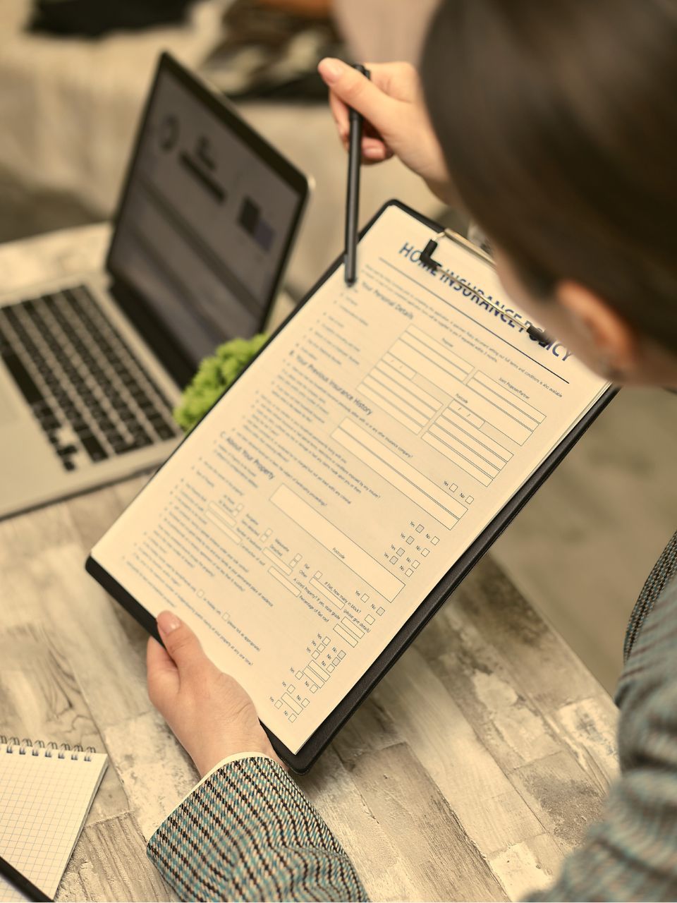 Person holding a clipboard with a form, pointing with a pen, laptop visible on a table.